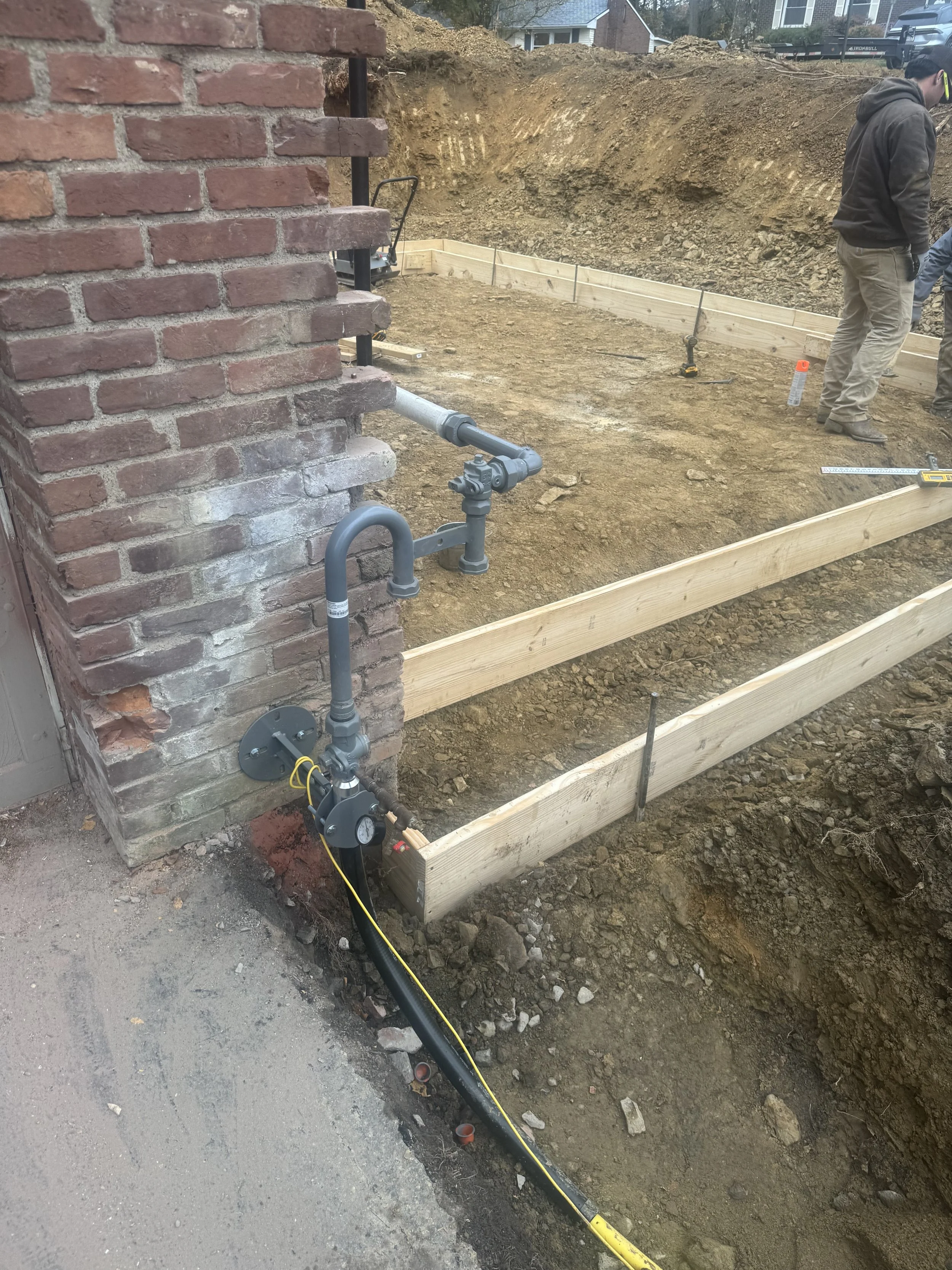 Construction site showing plumbing infrastructure near a brick wall, with wooden framing for a foundation or sidewalk, and workers in the background.