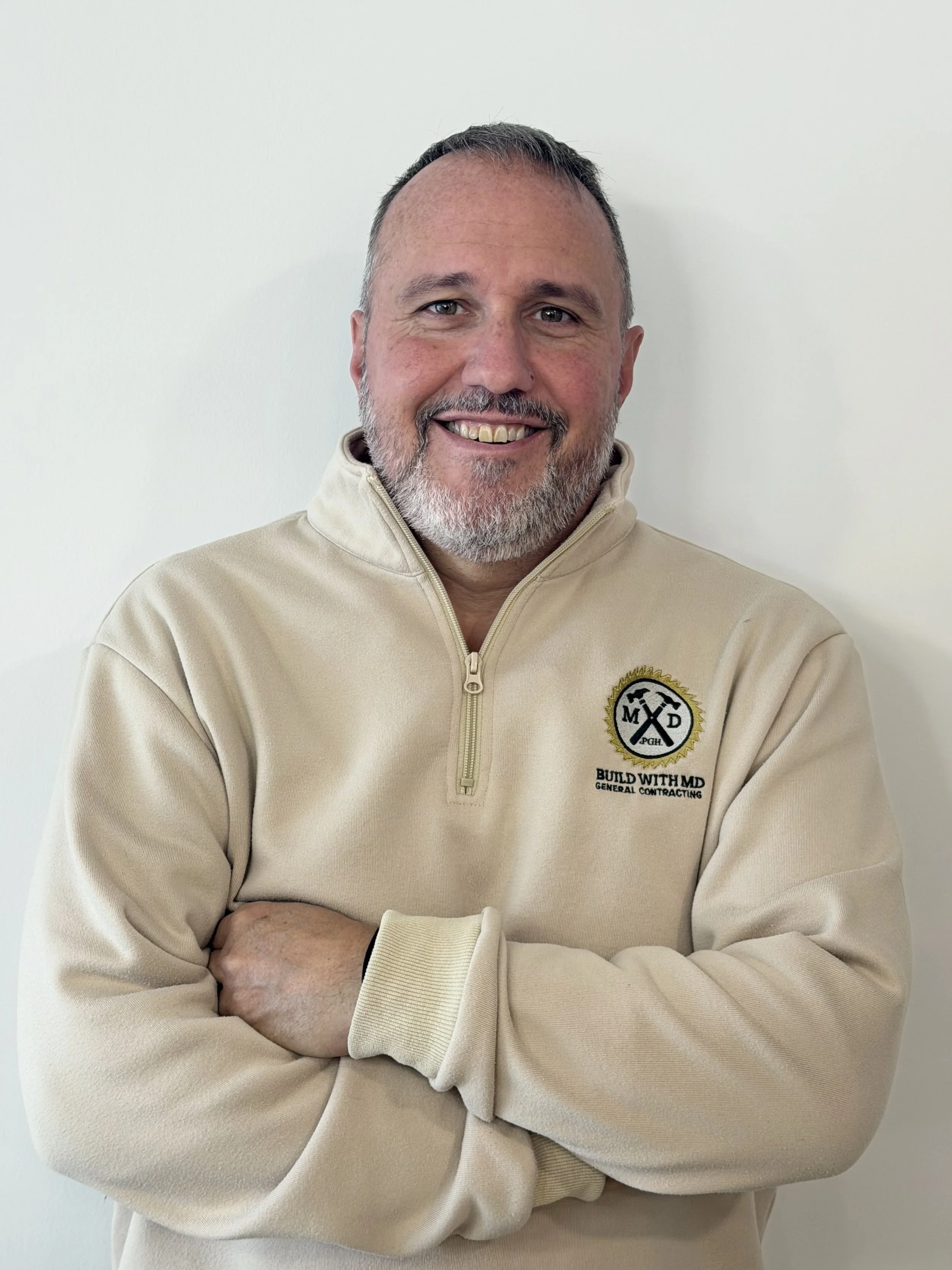A middle-aged man with a beard and short hair, smiling and crossing his arms, wearing a beige quarter-zip sweatshirt with a logo that says "Build With M.D. General Contracting".