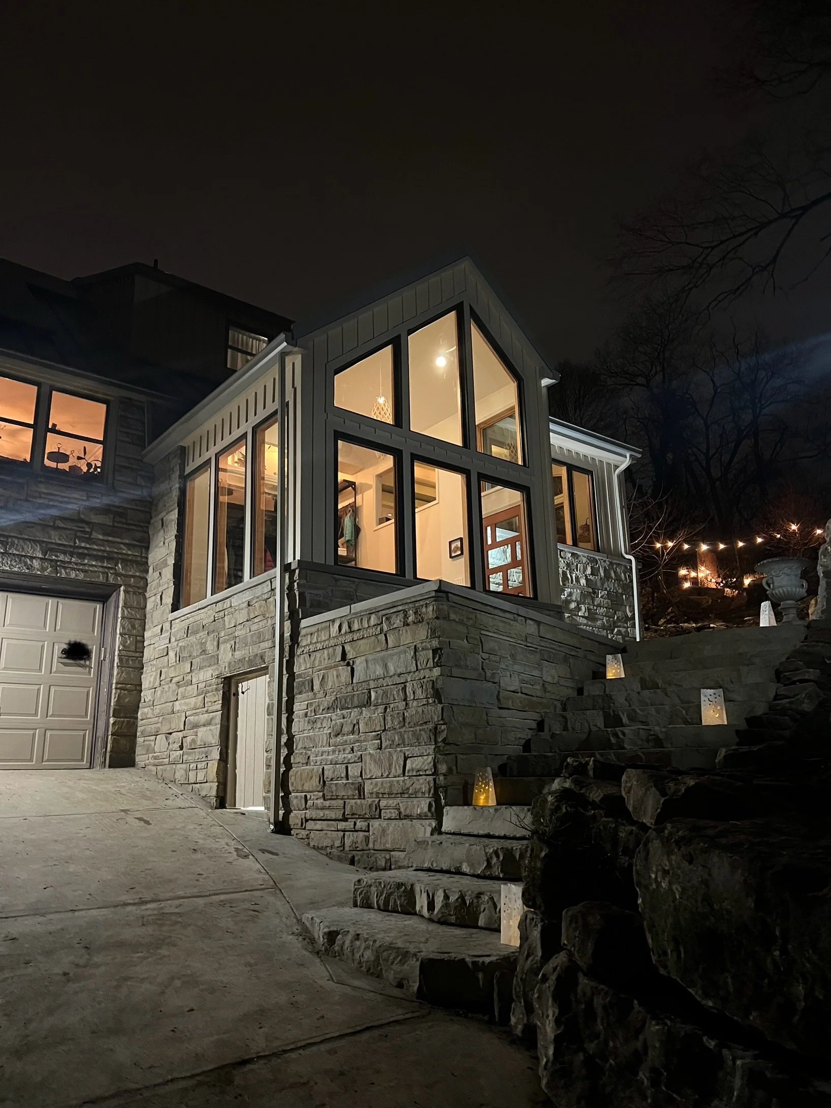 Nighttime view of a house with large illuminated windows and stone steps with lanterns leading up to it.