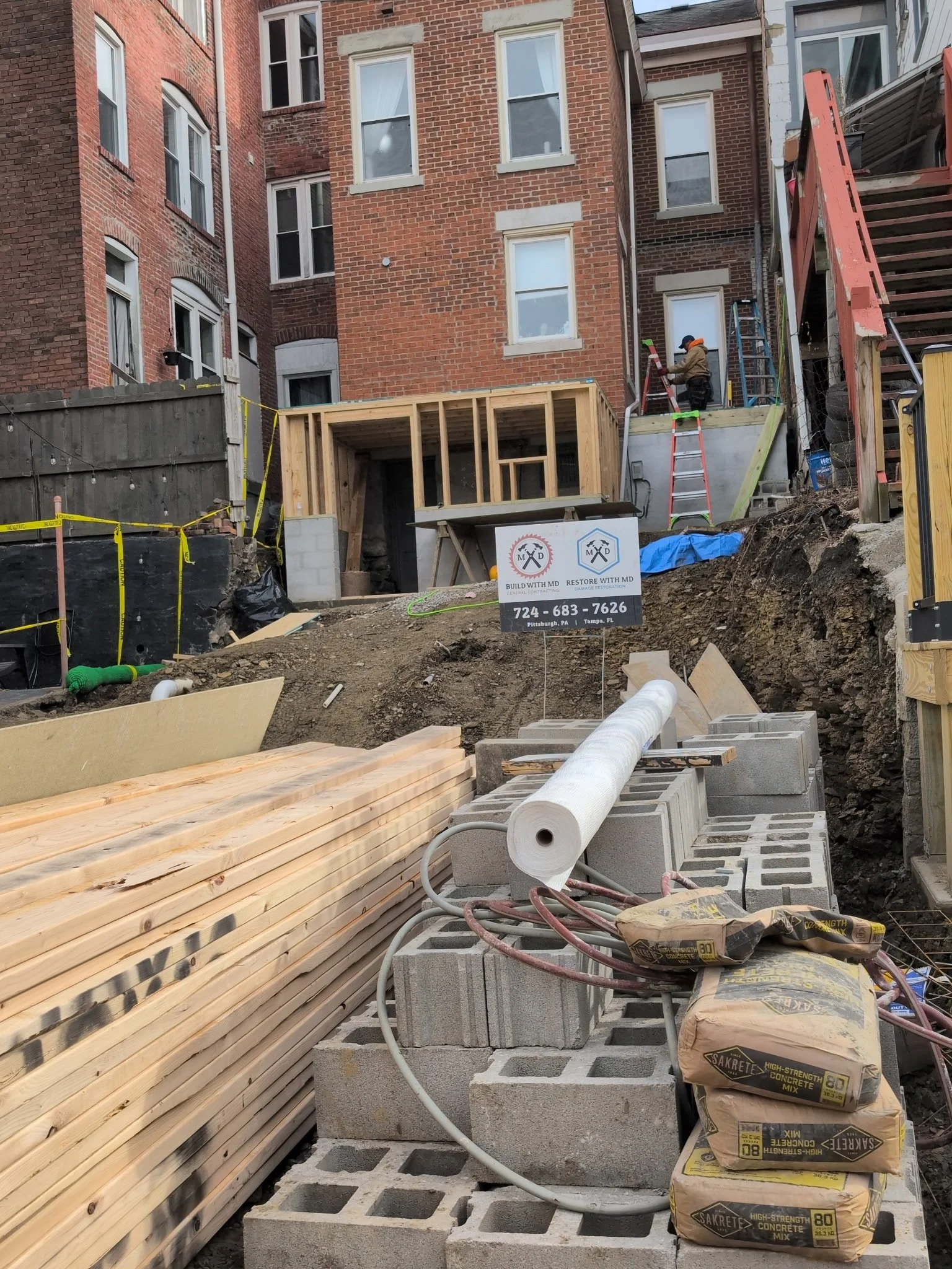 Construction site with a partially built wooden structure, piles of bricks, and construction workers working on an elevated platform in an urban area with brick buildings.