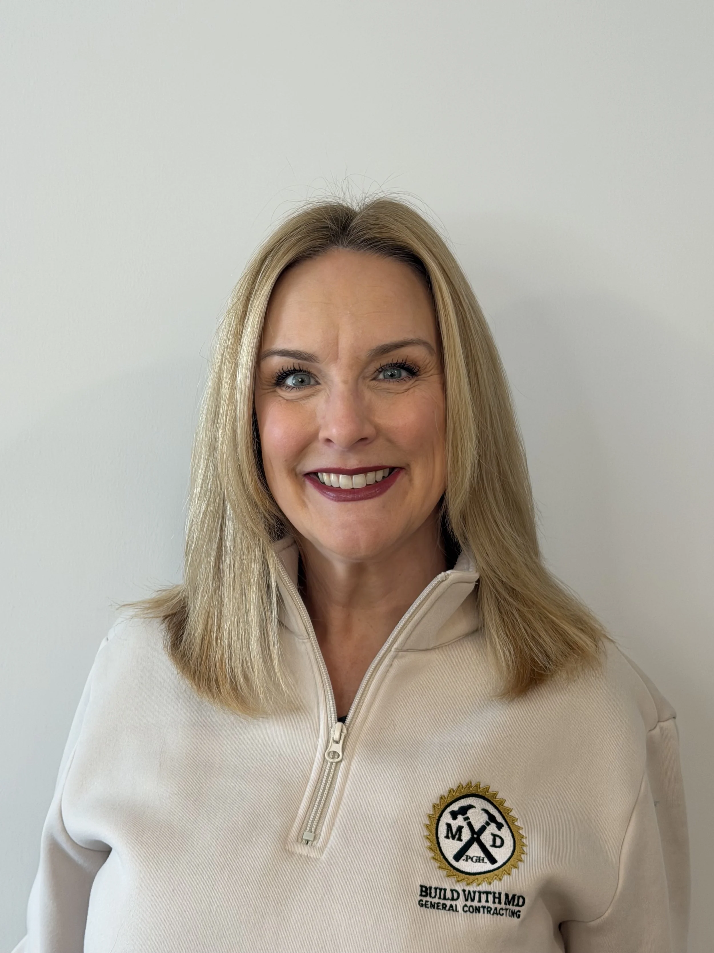 A woman with blonde hair smiling, wearing a beige sweatshirt with a logo and the text 'Build With MD General Contracting' on the chest, standing against a plain white wall.