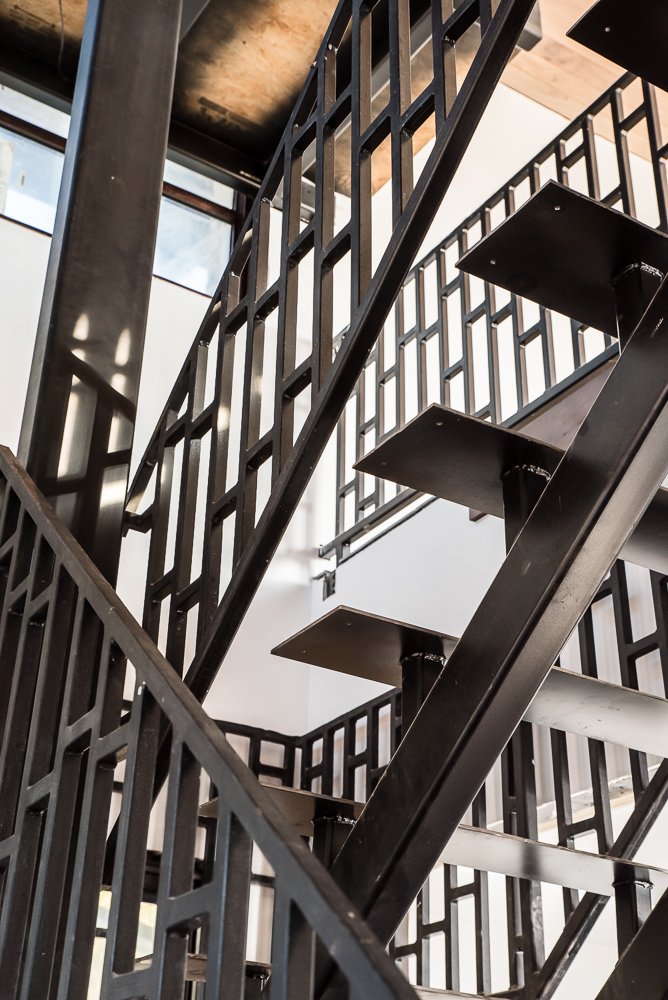 Interior view of a modern black metal staircase with angular steps and geometric railing design, in a building with large windows and wooden ceiling.