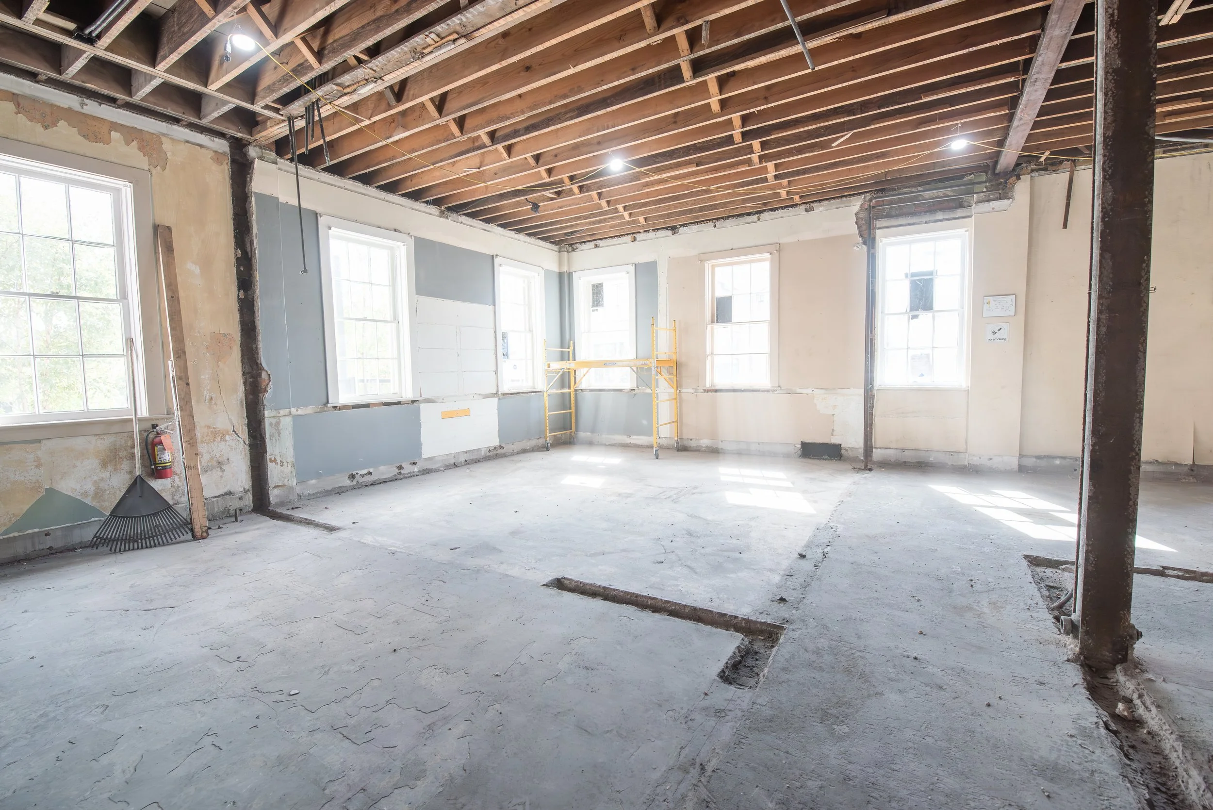 Empty room under renovation with exposed wooden ceiling beams, unpainted walls, and construction debris on the floor. There are multiple large windows allowing natural light in, and a yellow scaffolding in the corner.