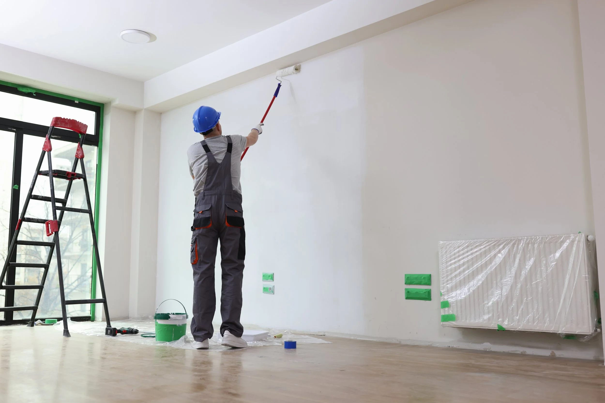 A construction worker wearing a blue helmet and gray overalls painting a white wall with a roller brush inside a room. There's a ladder and a collection of painting tools and supplies on the floor.