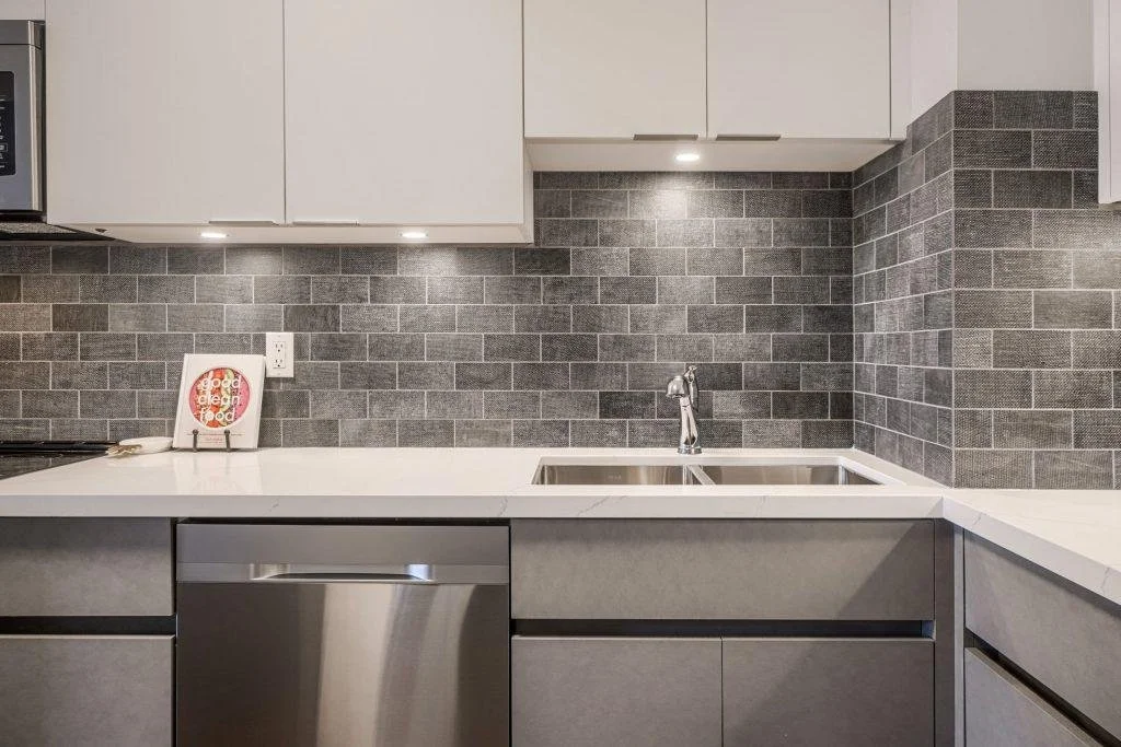 Modern kitchen with white upper cabinets, gray lower cabinets, a stainless steel dishwasher, a double sink, and a gray brick backsplash.