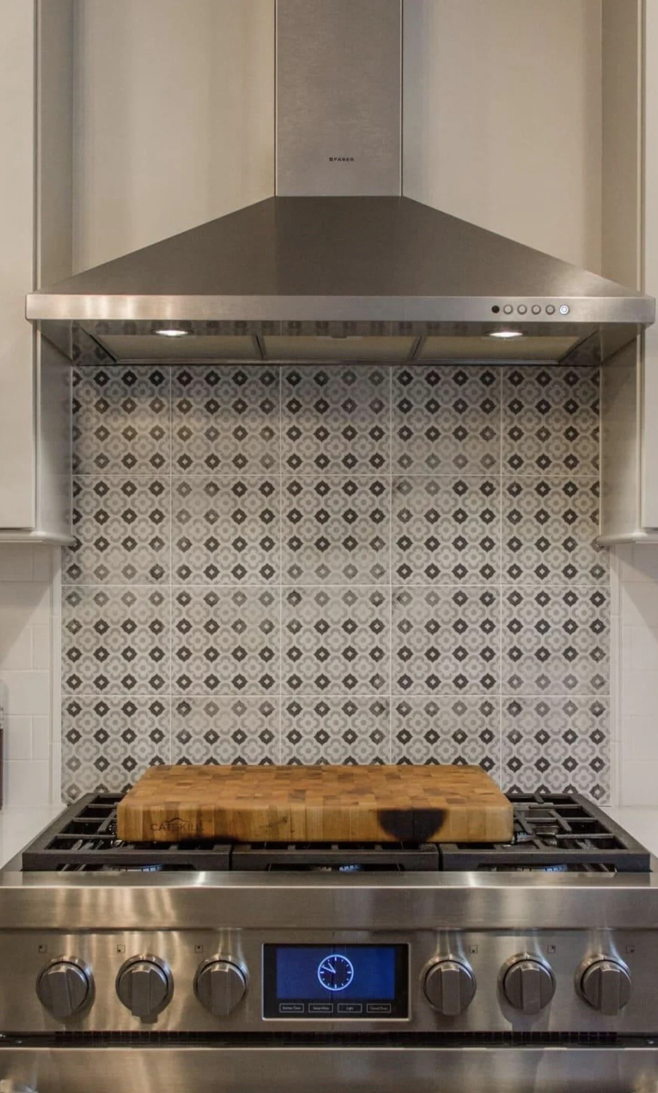 Stainless steel gas stove with six control knobs, digital display, wooden cutting board on burners, patterned tile backsplash, and range hood above.