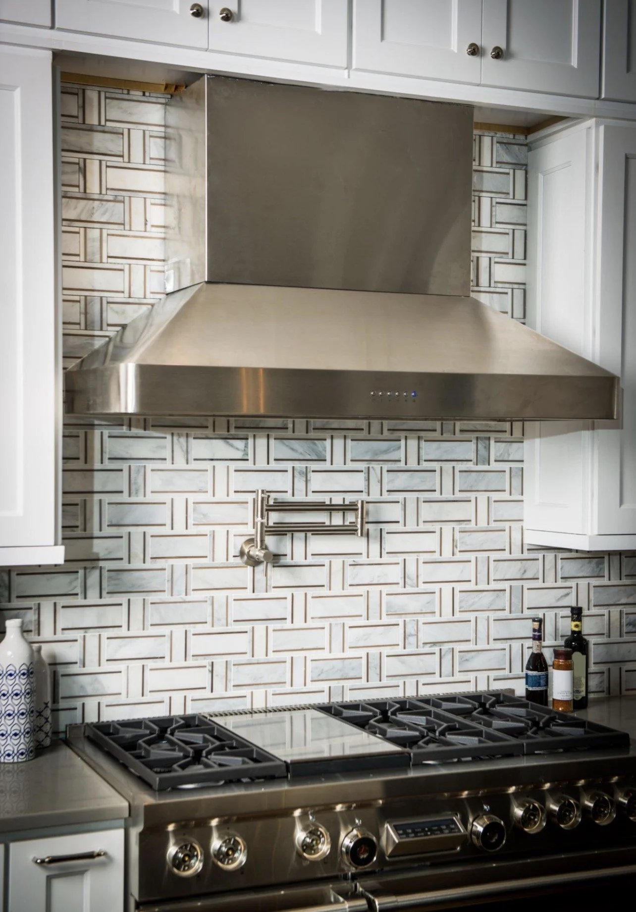 Kitchen with a stainless steel gas stove and a gray backsplash with a brick-like tile pattern. There is a vent hood above the stove, white cabinetry, and some bottles on the counter.