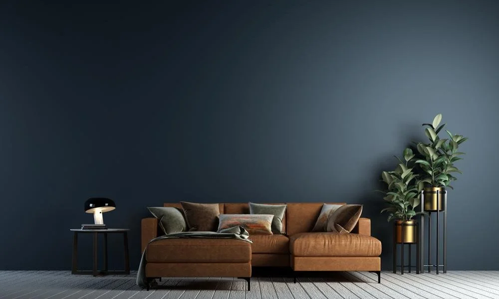 Modern interior with a brown sofa, multiple pillows, a small side table with a lamp, and tall green plants in gold planters against a dark blue wall.