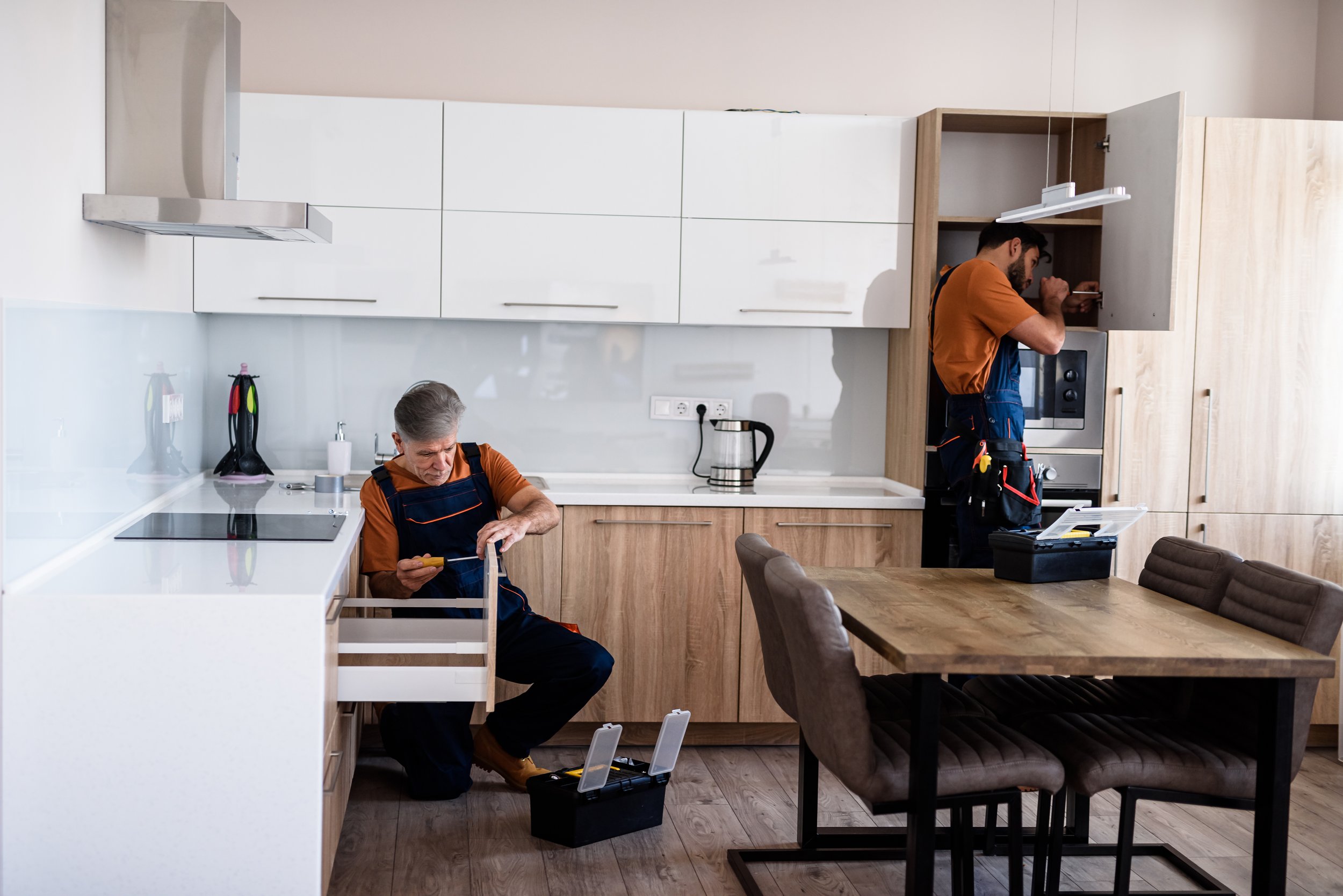 Two repairmen working in a modern kitchen, one kneeling and examining a drawer, the other standing near a cabinet with tools and equipment.