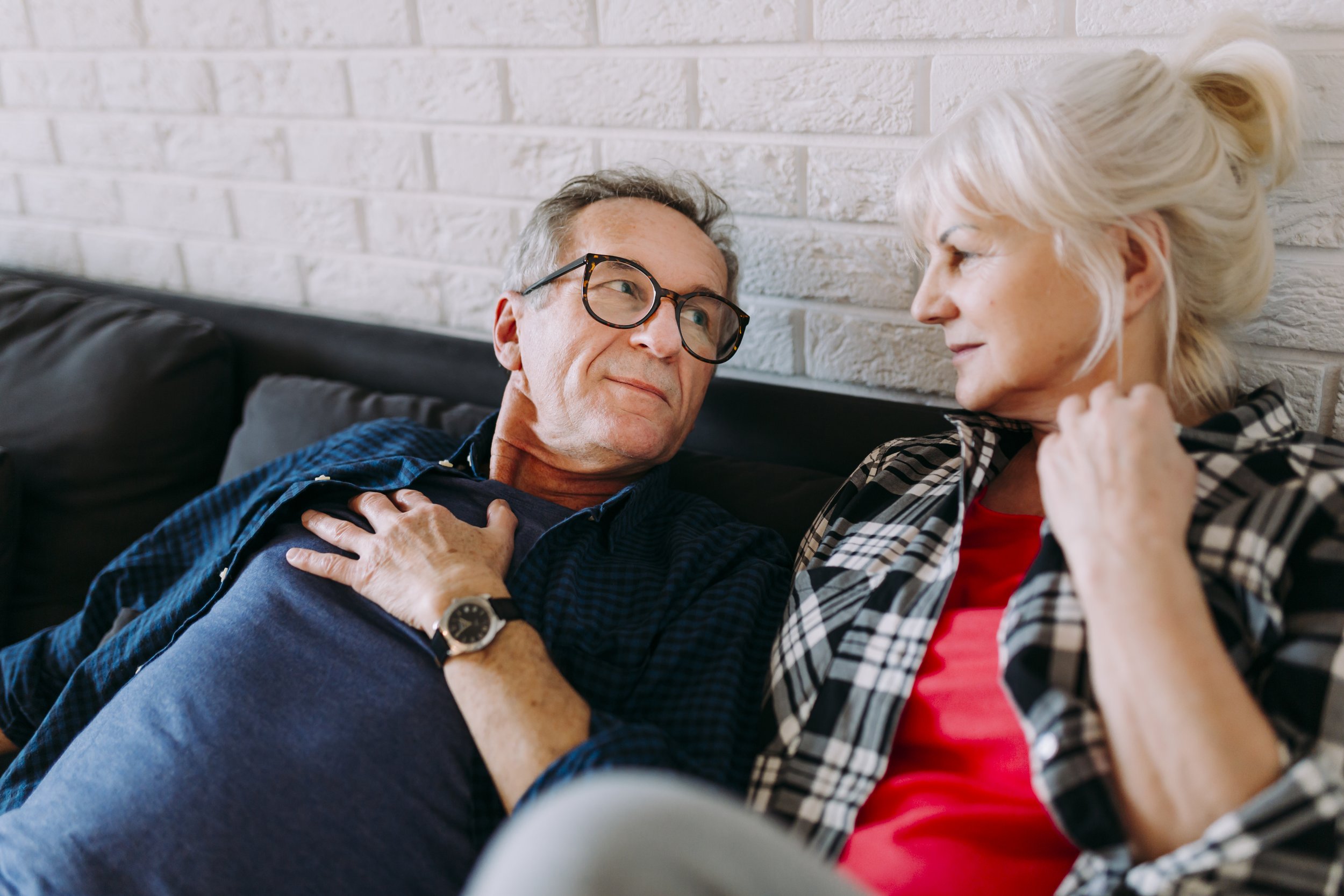A middle-aged man with glasses and a woman with blonde hair sitting on a couch, sharing a tender moment as they look at each other.
