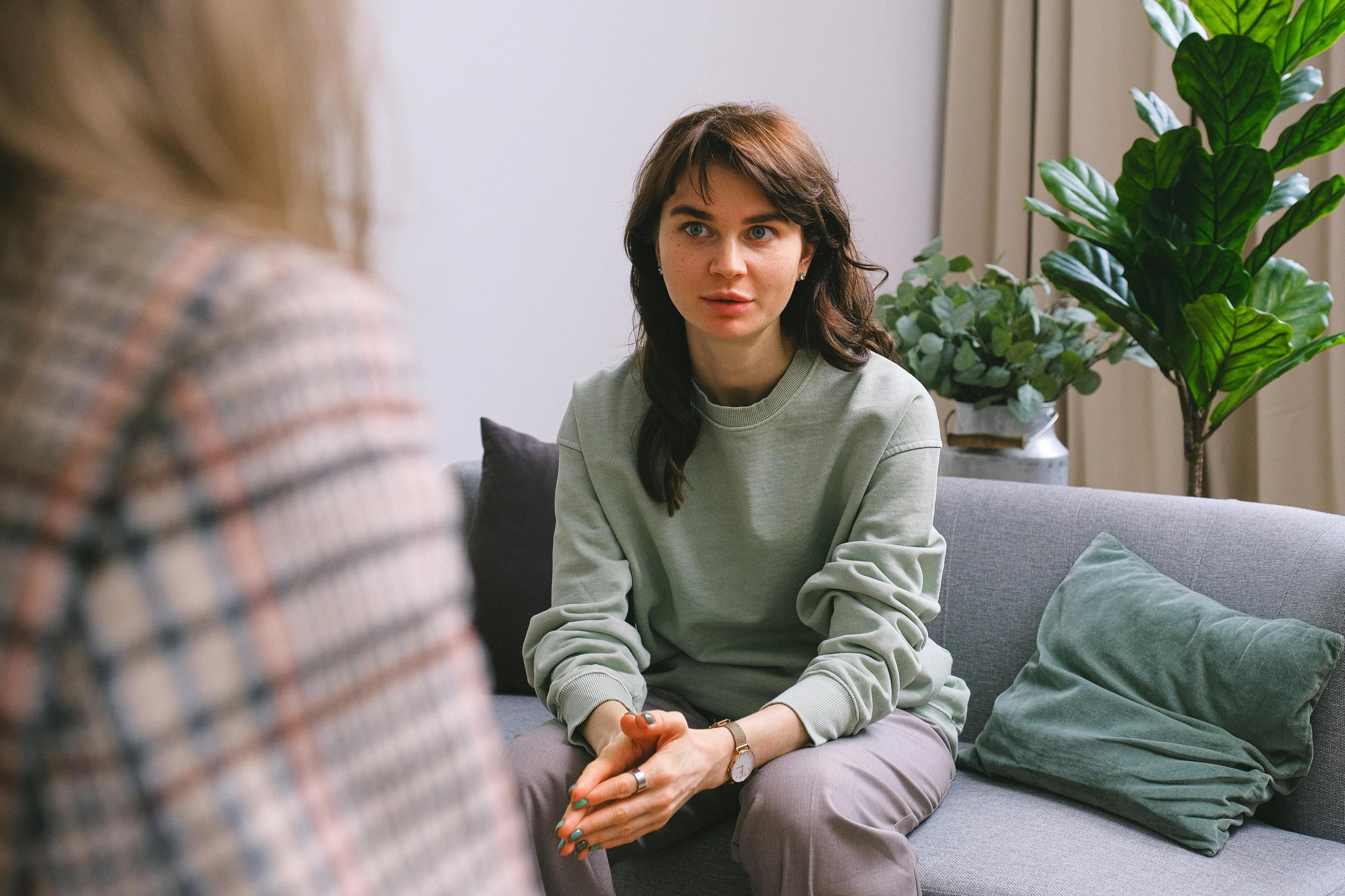 A woman with brown hair and blue eyes sitting on a gray couch in a room with a potted plant in the background, engaged in conversation with another person.