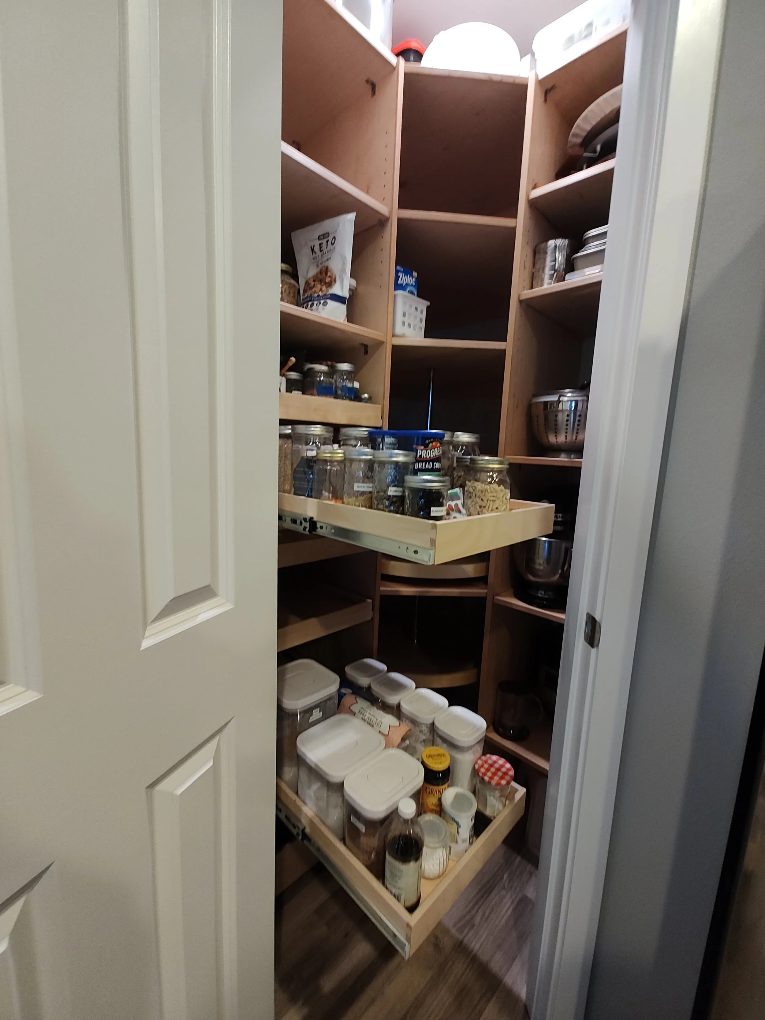 A pantry with wooden shelves filled with jars, containers, and food items, with some contents visible like spices, snacks, and baking supplies.