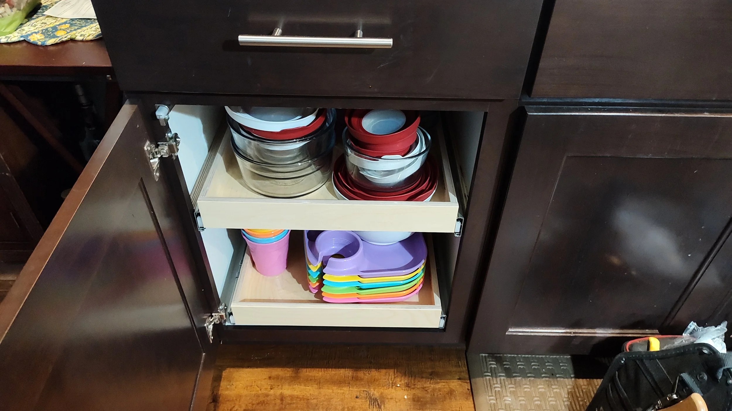 Open kitchen cabinet with two wooden pull-out shelves containing glass bowls, plastic cups, and colorful plastic trays.