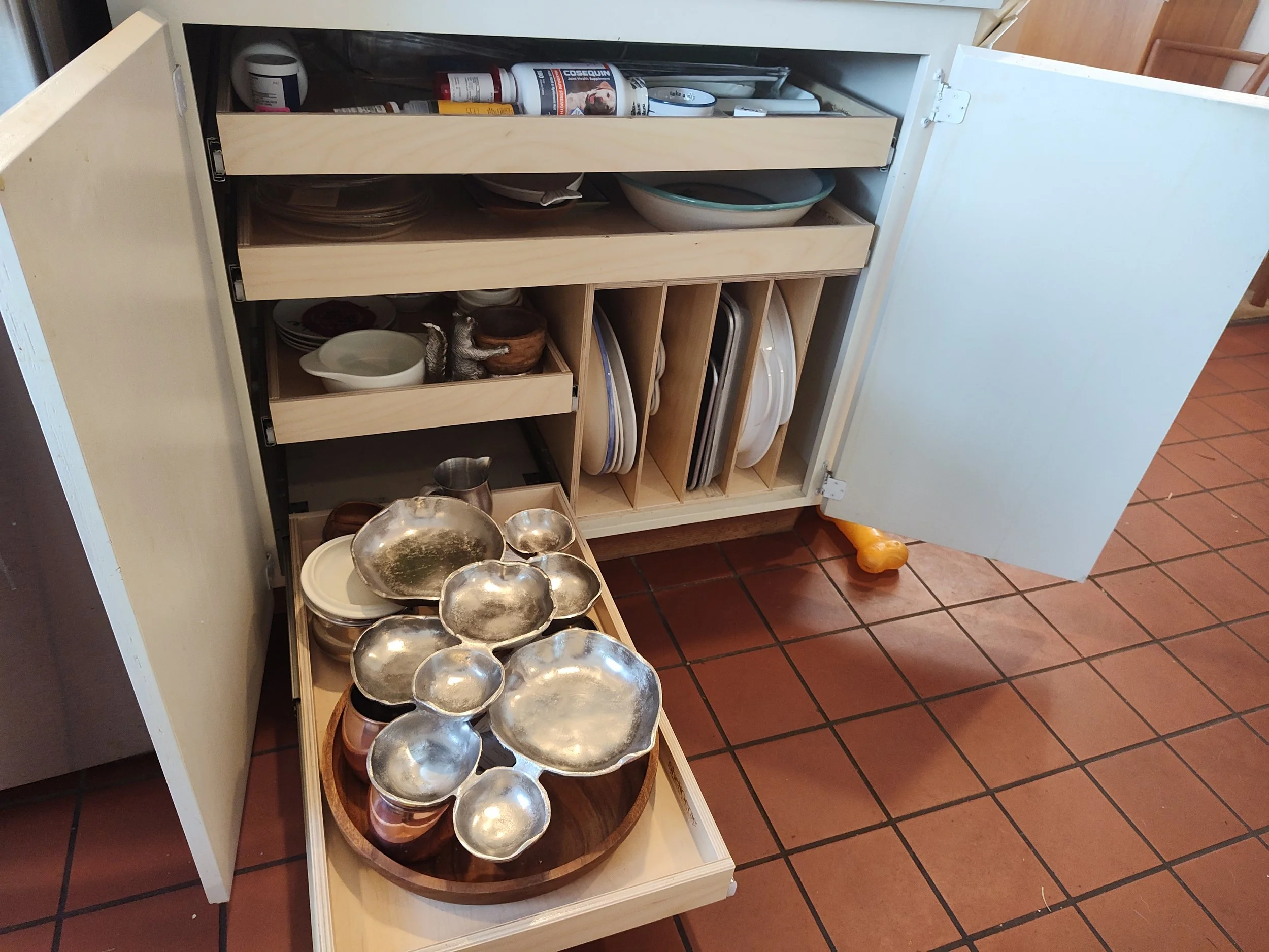 Open kitchen cabinet with dishes, bowls, and utensils; some dishes are stacked or in compartments, with metal and ceramic bowls on a tray in front.