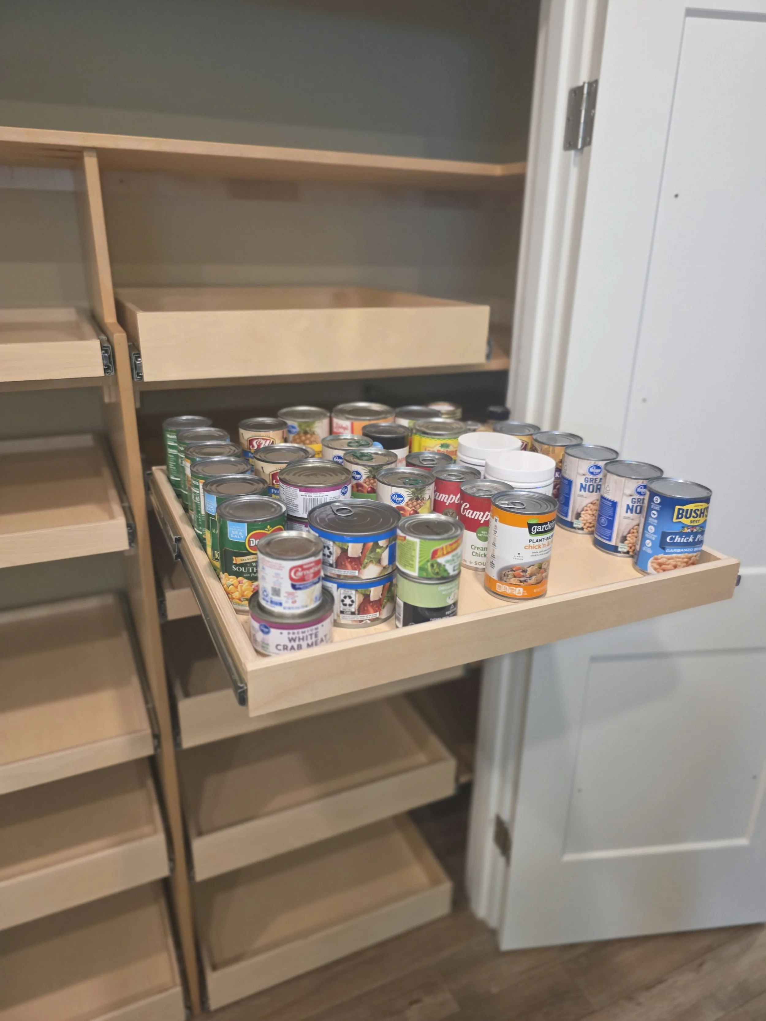 An open wooden pull-out shelf in a kitchen cabinet filled with canned foods, including soup, crab meat, and beans.