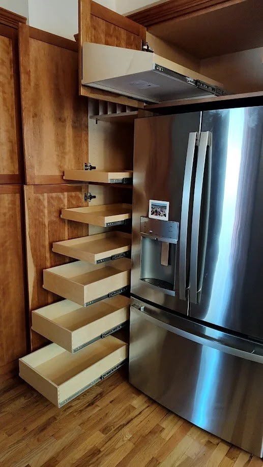 A kitchen with wooden cabinets and a stainless steel refrigerator. The refrigerator has its freezer door on the bottom and a water and ice dispenser. To the left of the refrigerator, there are several beige pull-out drawers or shelves extending from 