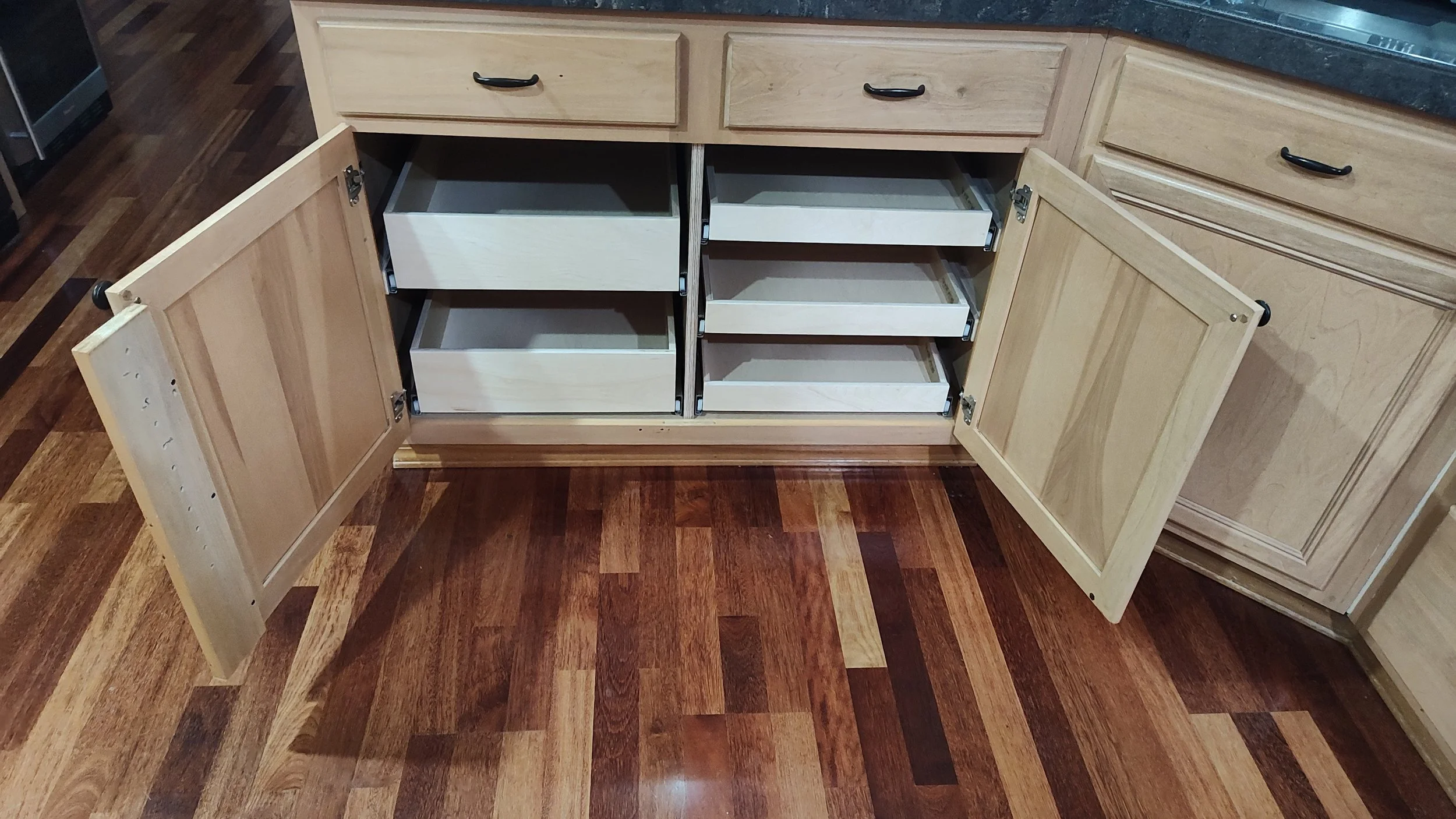 Open kitchen cabinet with several empty white drawers, in a wooden cabinet under a black countertop, with a hardwood floor.
