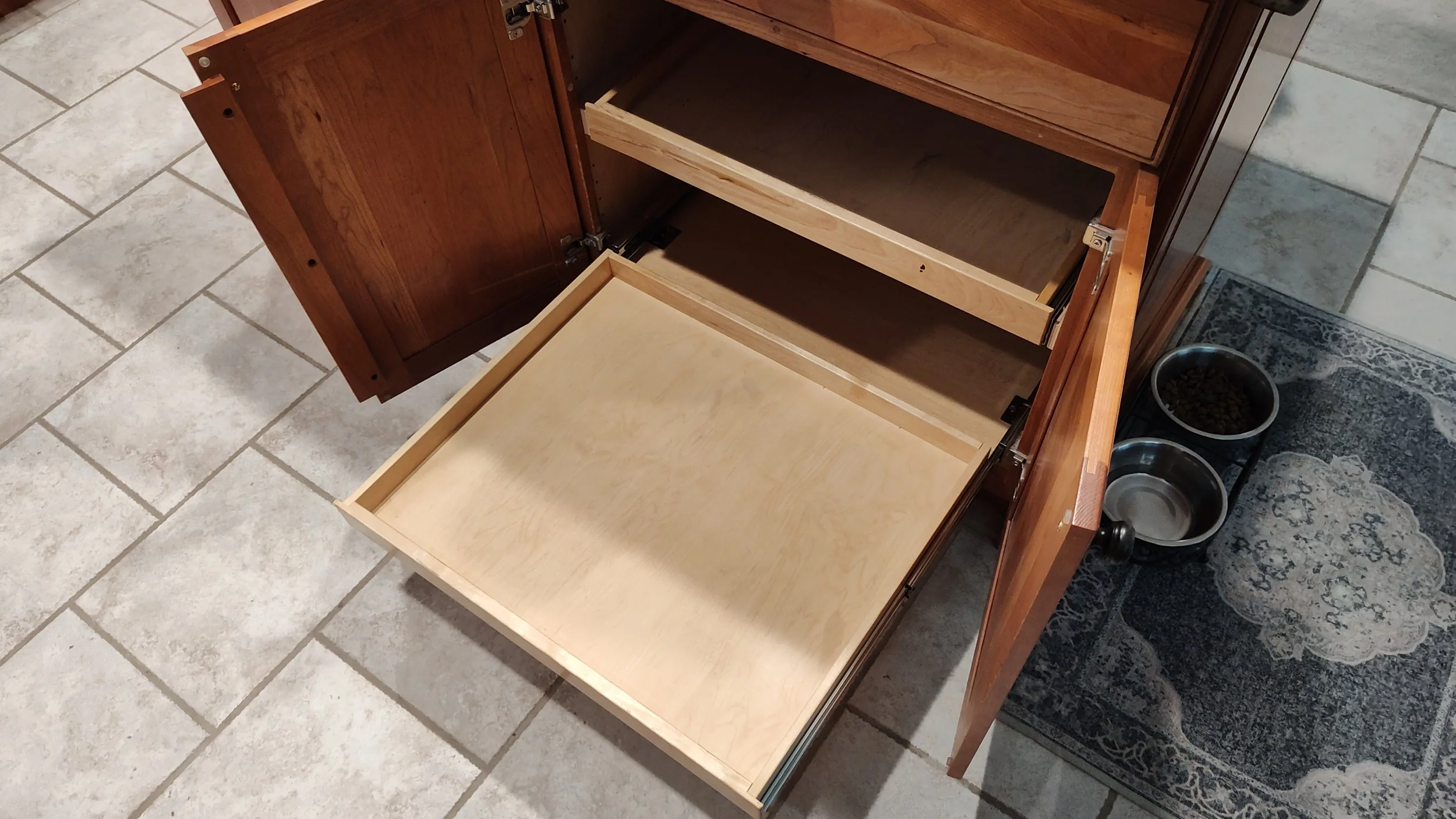 Open wooden kitchen cabinet with empty drawers, situated on tiled floor next to pet bowls on a small rug.