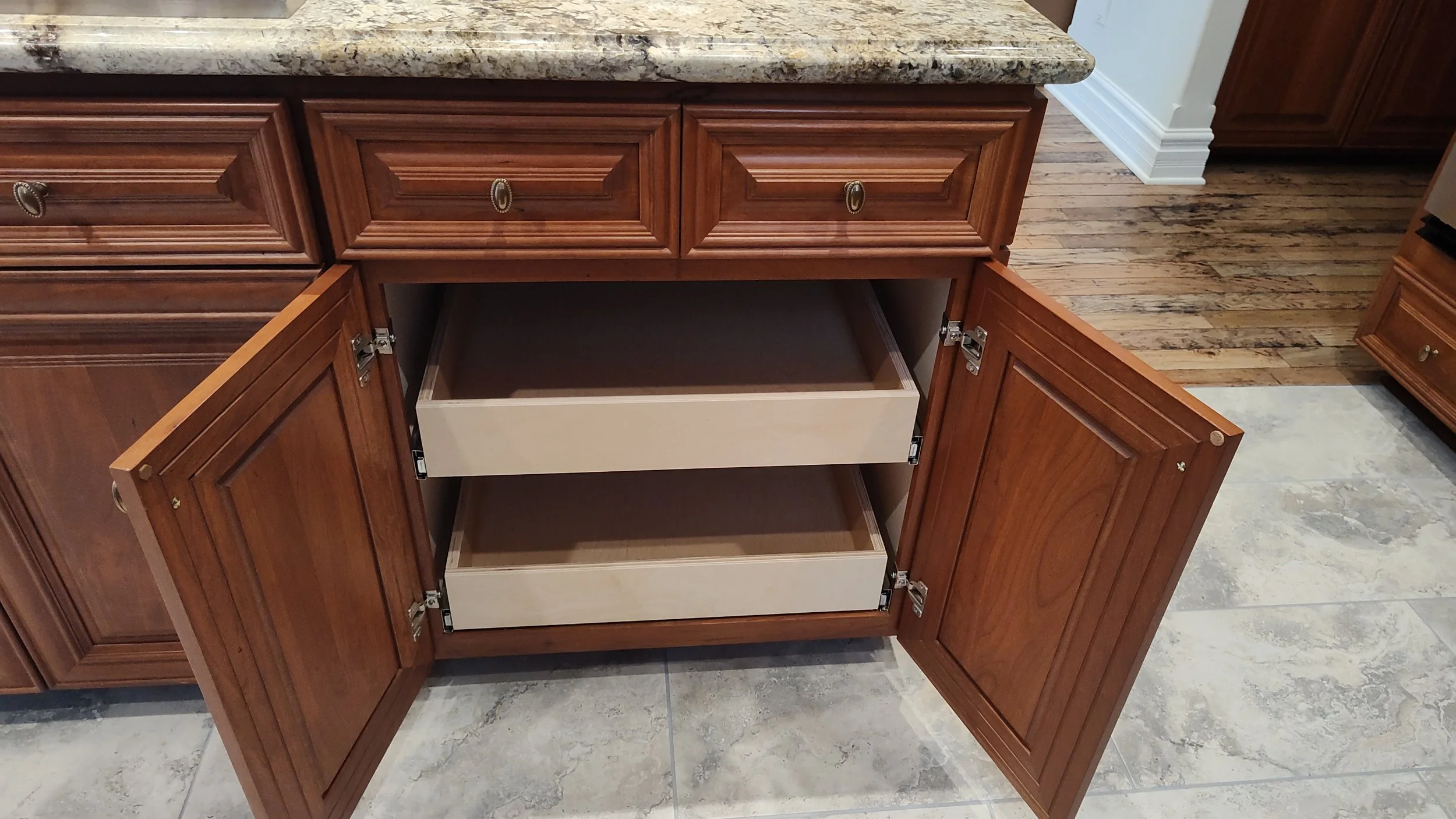 Open kitchen cabinet with two empty shelves and brown wooden doors in a kitchen with a granite countertop and tiled floor.