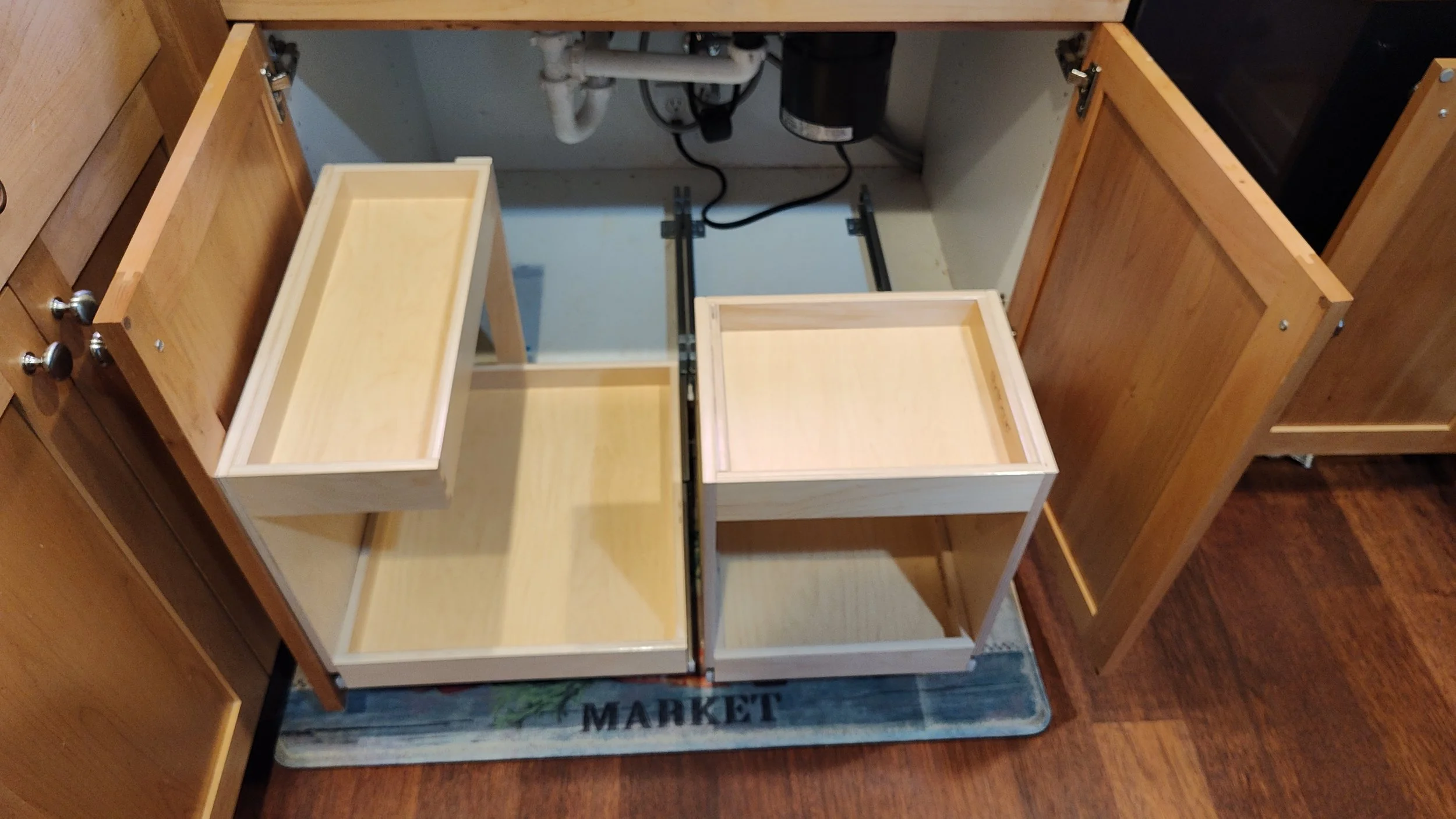Unassembled wooden drawers inside a kitchen cabinet with open doors, sitting on a mat on a hardwood floor.