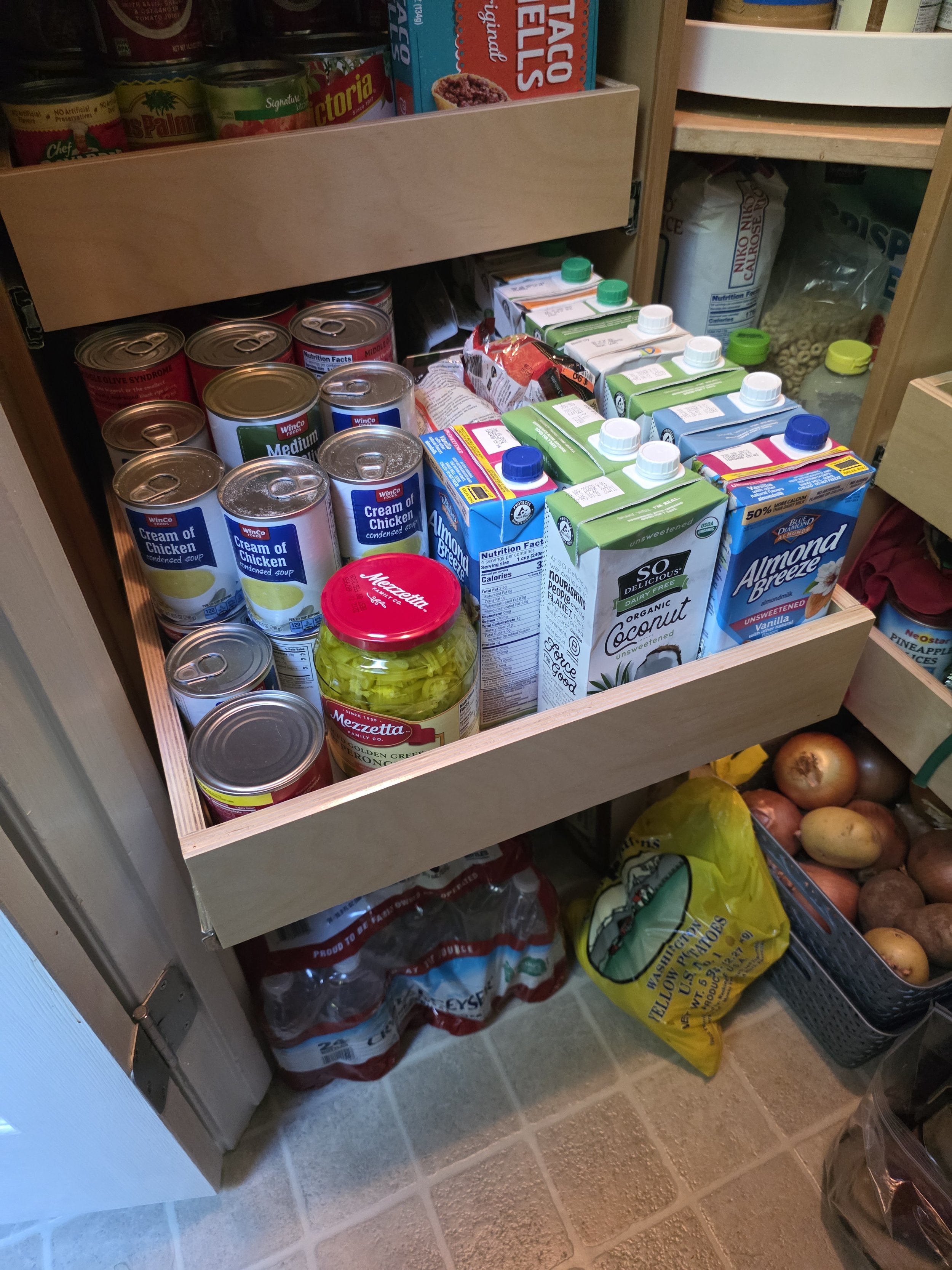 Open pantry drawer containing cans of soup, almond milk, coconut milk, and a jar of pickles, with onions stored beneath and a bag of potatoes nearby.
