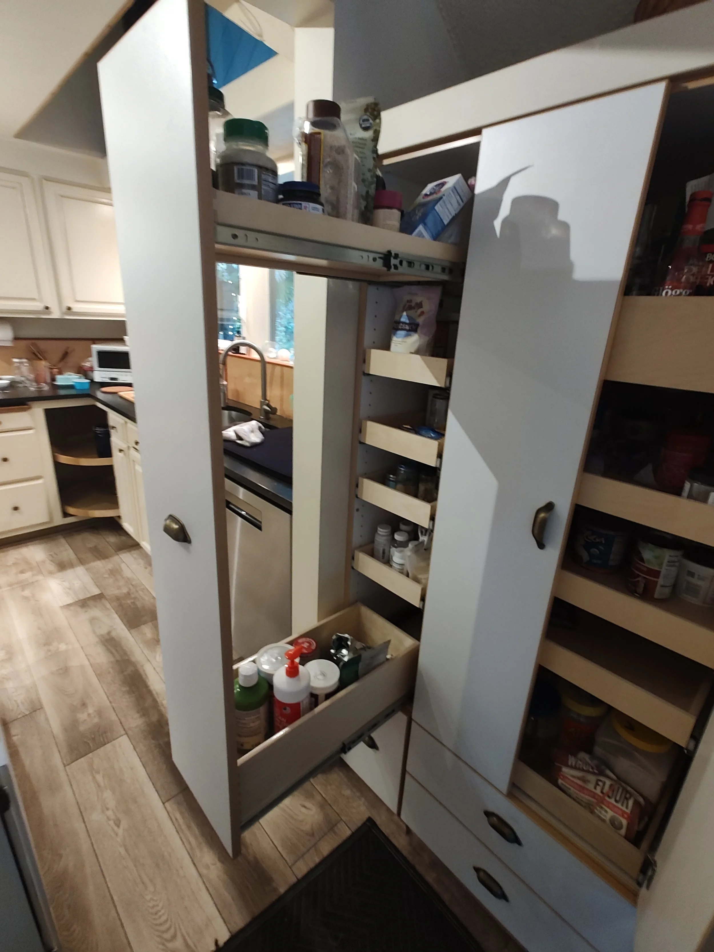 Open kitchen cabinet with various supplies including spices, bottles, and cleaning products, with a view of the kitchen and a window behind the sink.