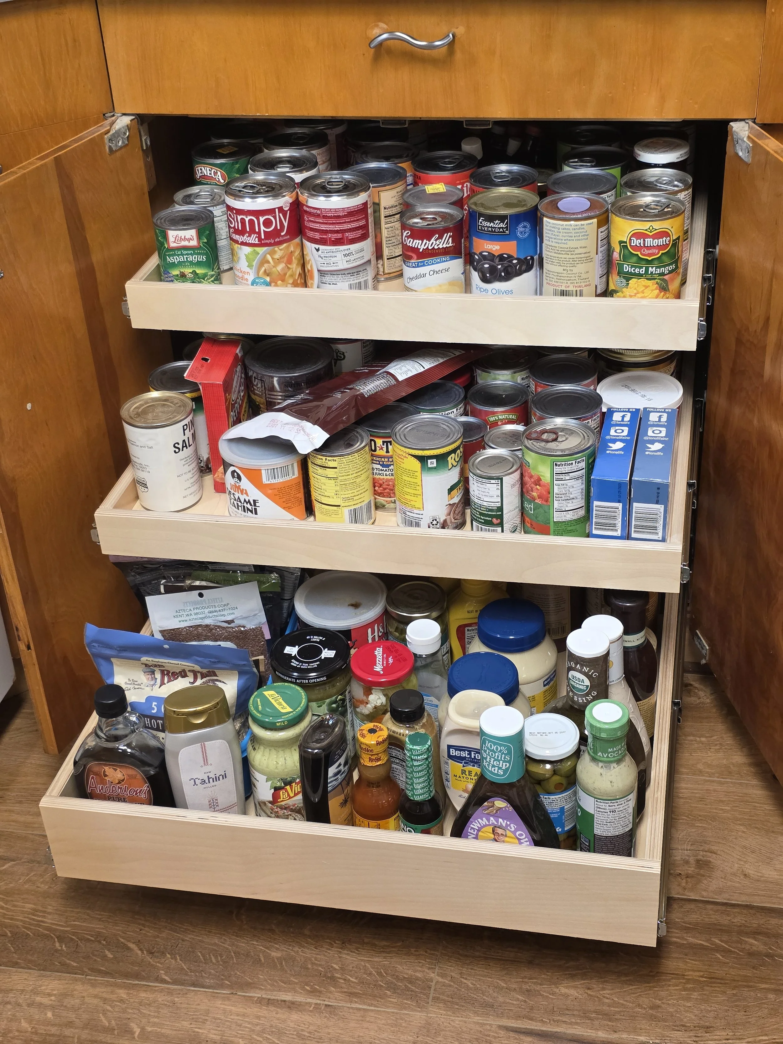 A kitchen cabinet with three shelves filled with canned goods, jars, bottles, and condiments.