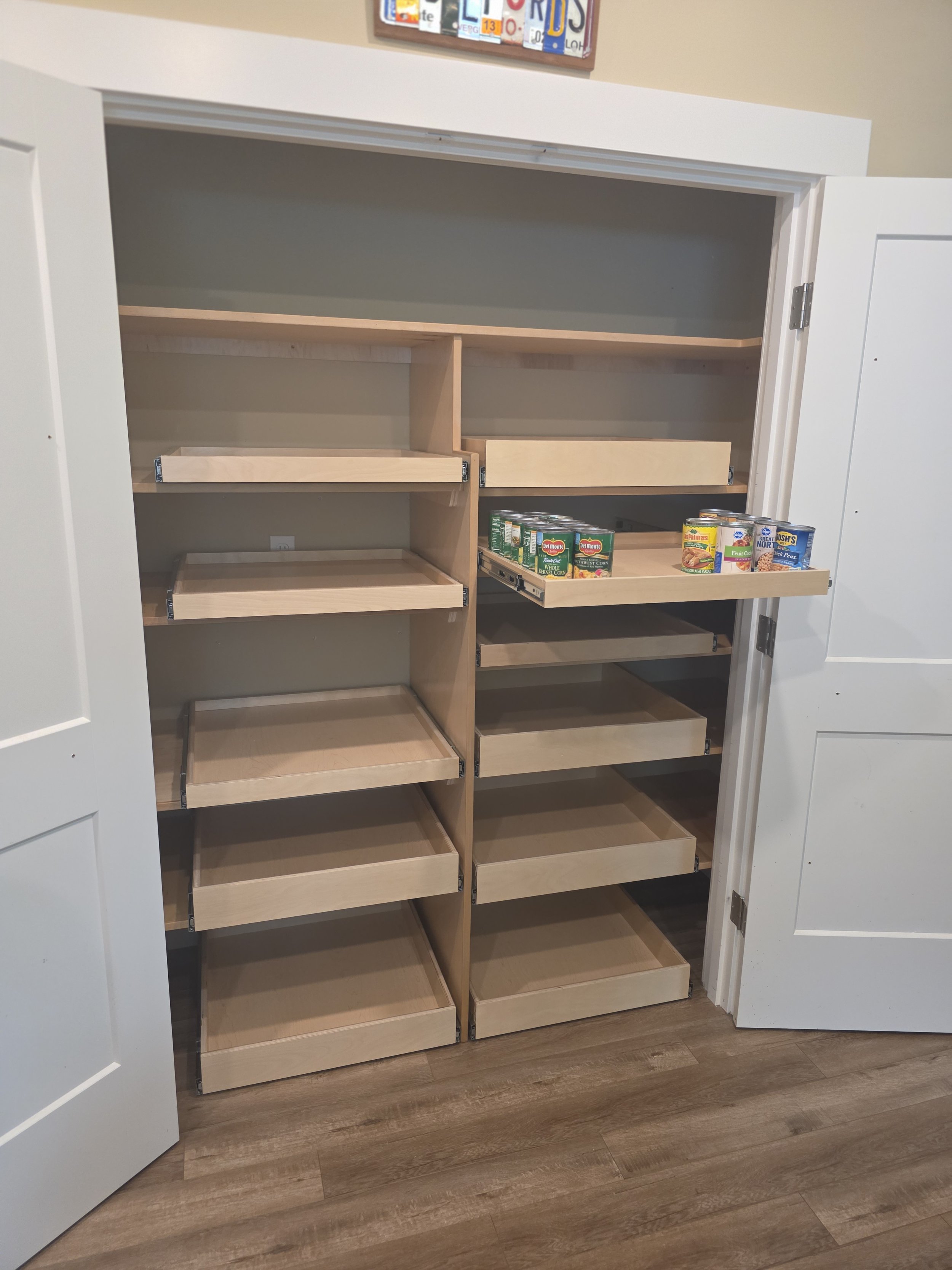 Empty wooden pantry with three shelves on the left and four on the right, partially filled with canned food items on the top shelf of the right side. White double doors open to reveal the pantry interior. A framed picture is visible above the pantry.