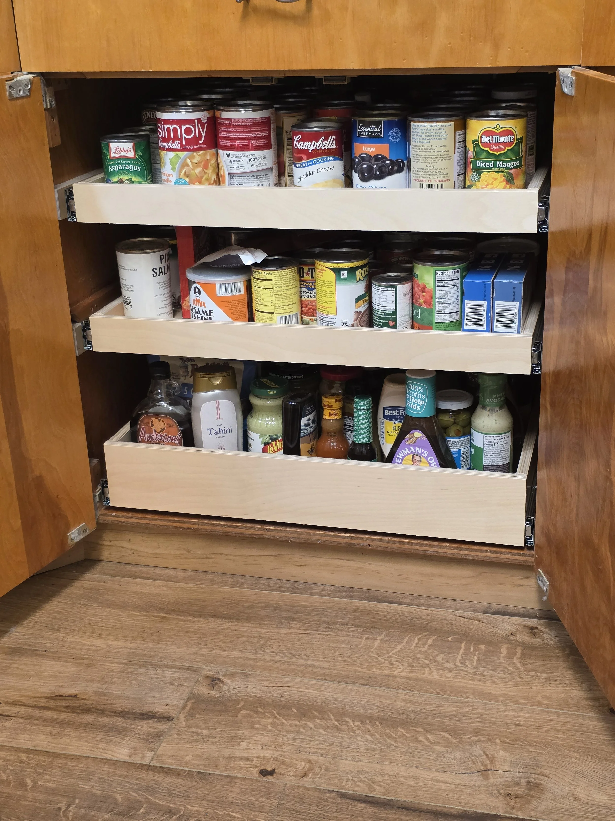 Under-cabinet kitchen pantry with three pull-out drawers filled with canned goods, condiments, and other food items.