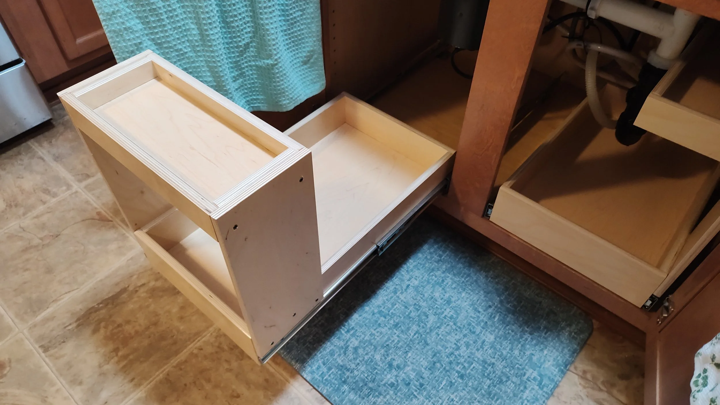 Unfinished wooden cabinet drawer slide-out section partially pulled out from a kitchen cabinet, with visible screws and structure, sitting on a tiled floor.