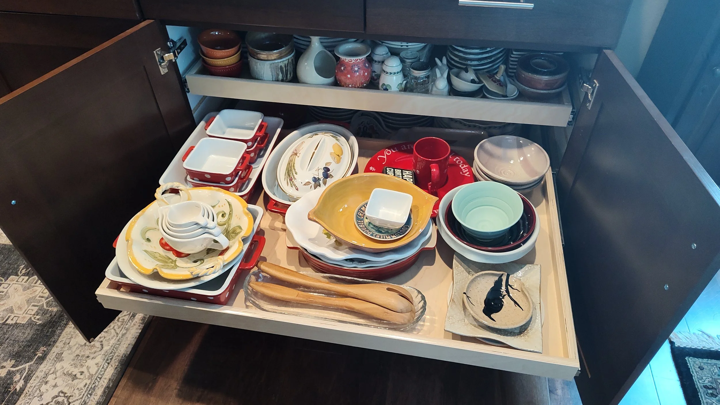 Open kitchen cabinet drawer filled with various dishes, bowls, and cups, including colorful ceramic and glassware items stacked and arranged inside.