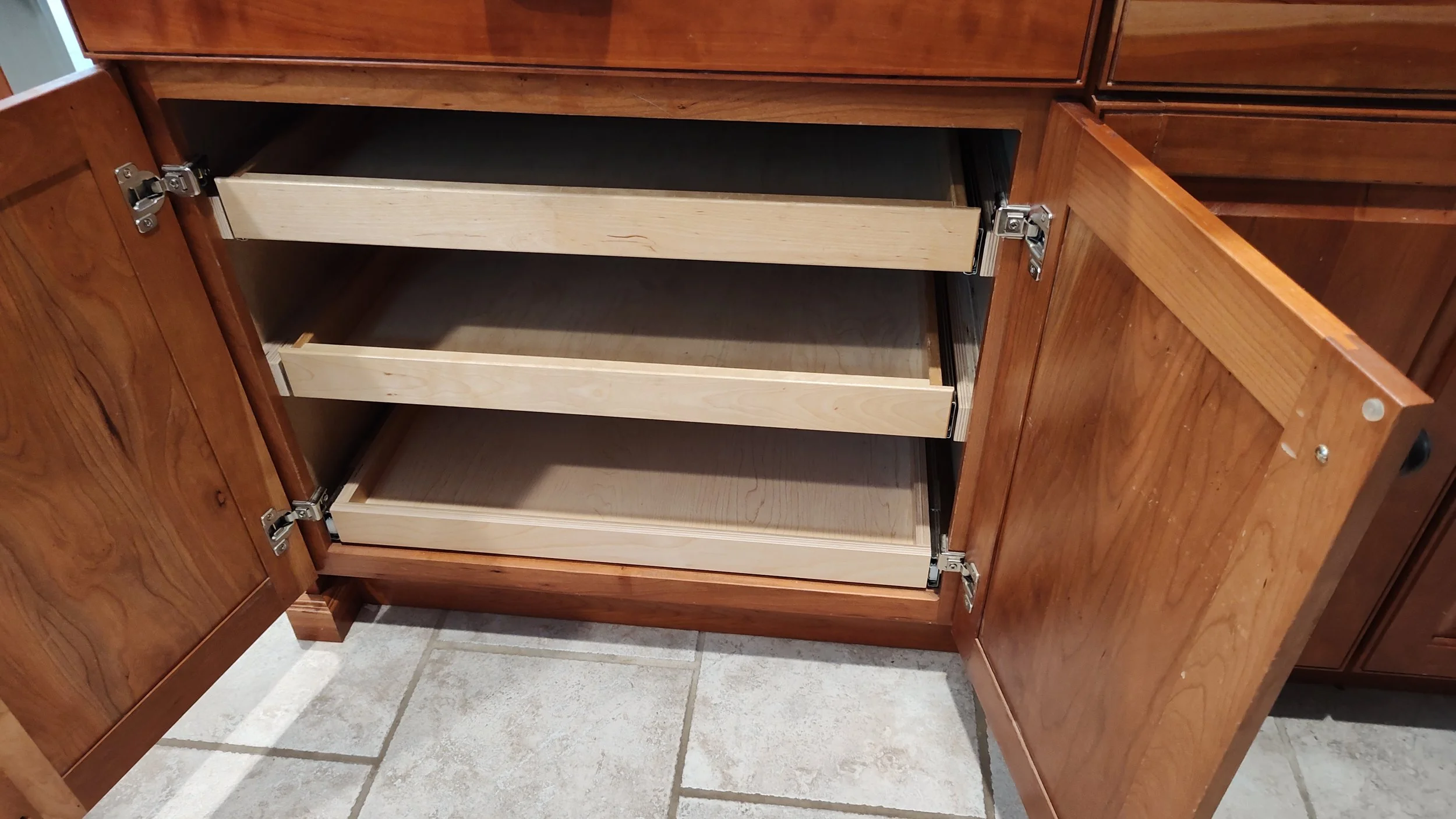Open wooden cabinet with three empty drawers, two of which are partially pulled out, showing the empty interior. The cabinet is on a tiled floor.