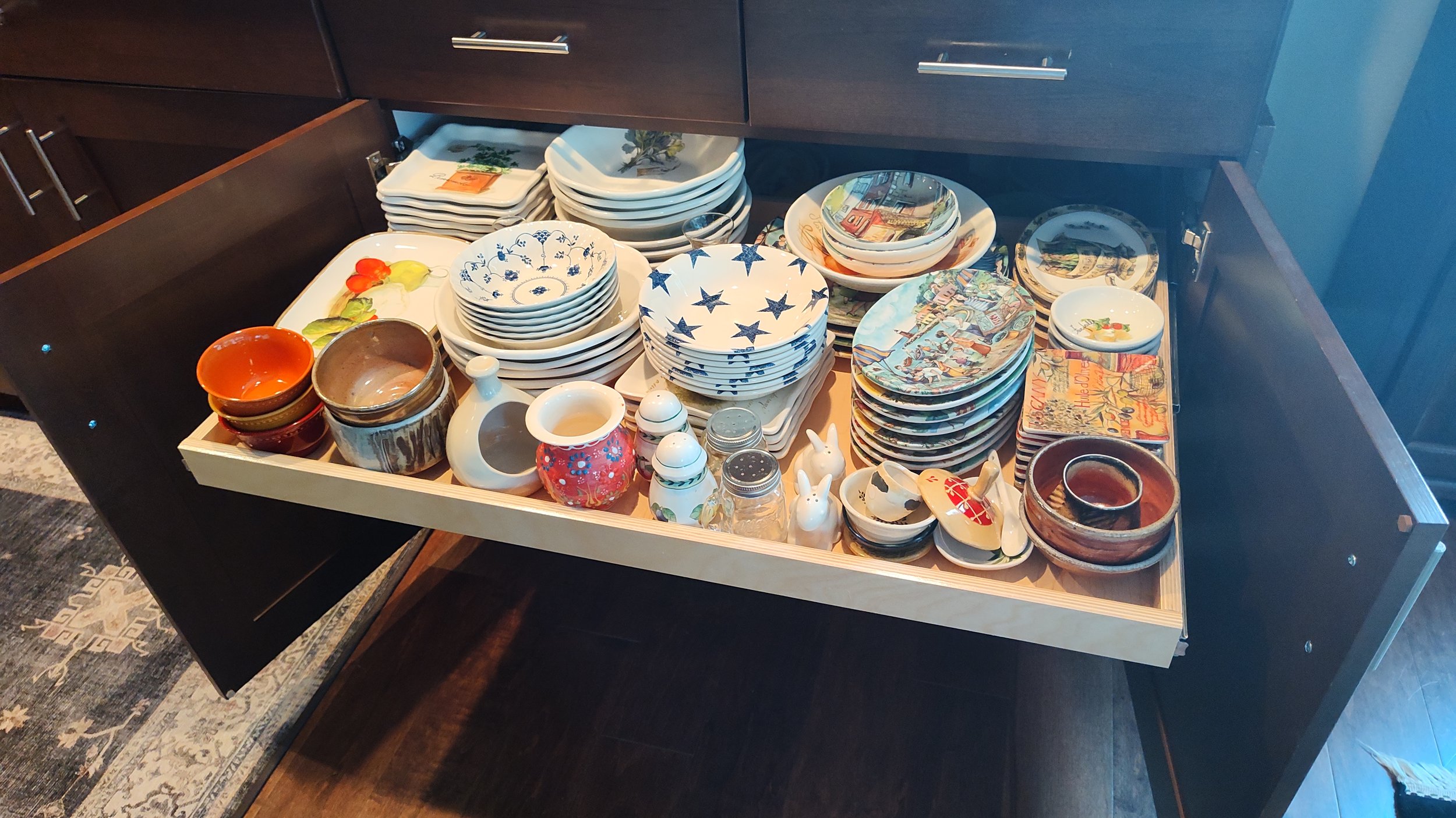 Open kitchen cabinet drawer filled with various decorative and colorful bowls, plates, and small kitchen accessories.