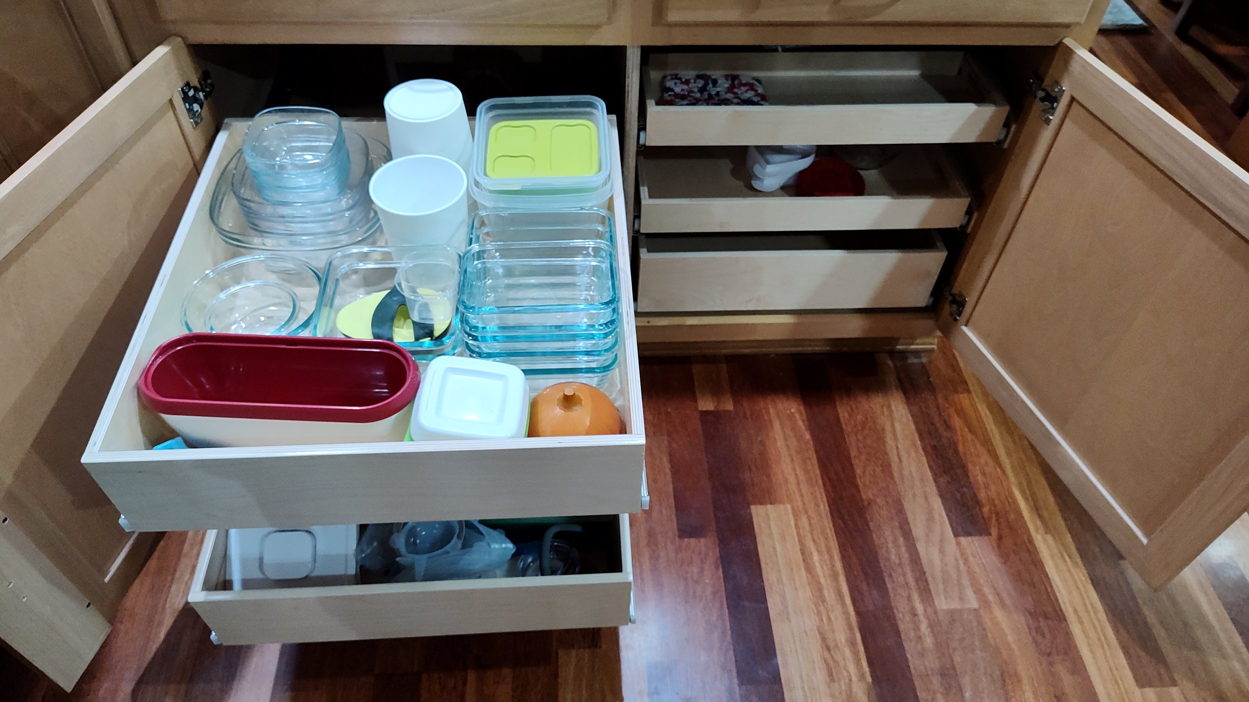 Open kitchen drawer with various glass dishes, bowls, and containers inside, and an orange plastic container on top, with cabinets and hardwood floor visible.