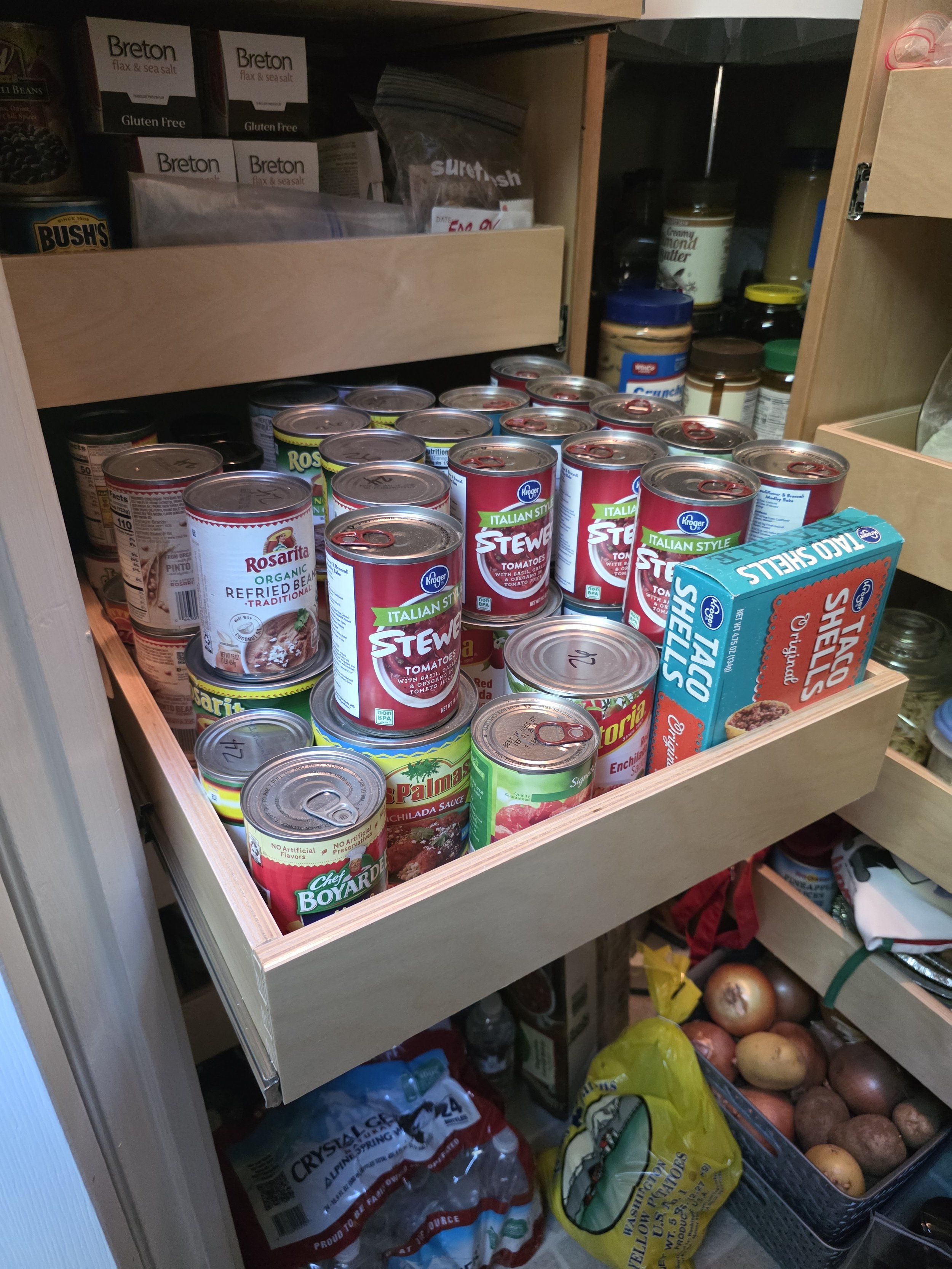 Open wooden pantry drawer filled with canned tomatoes, canned beans, and other canned goods, with shelves above containing boxes of gluten-free Breton sea salt and flax seeds, and various jars and food items.