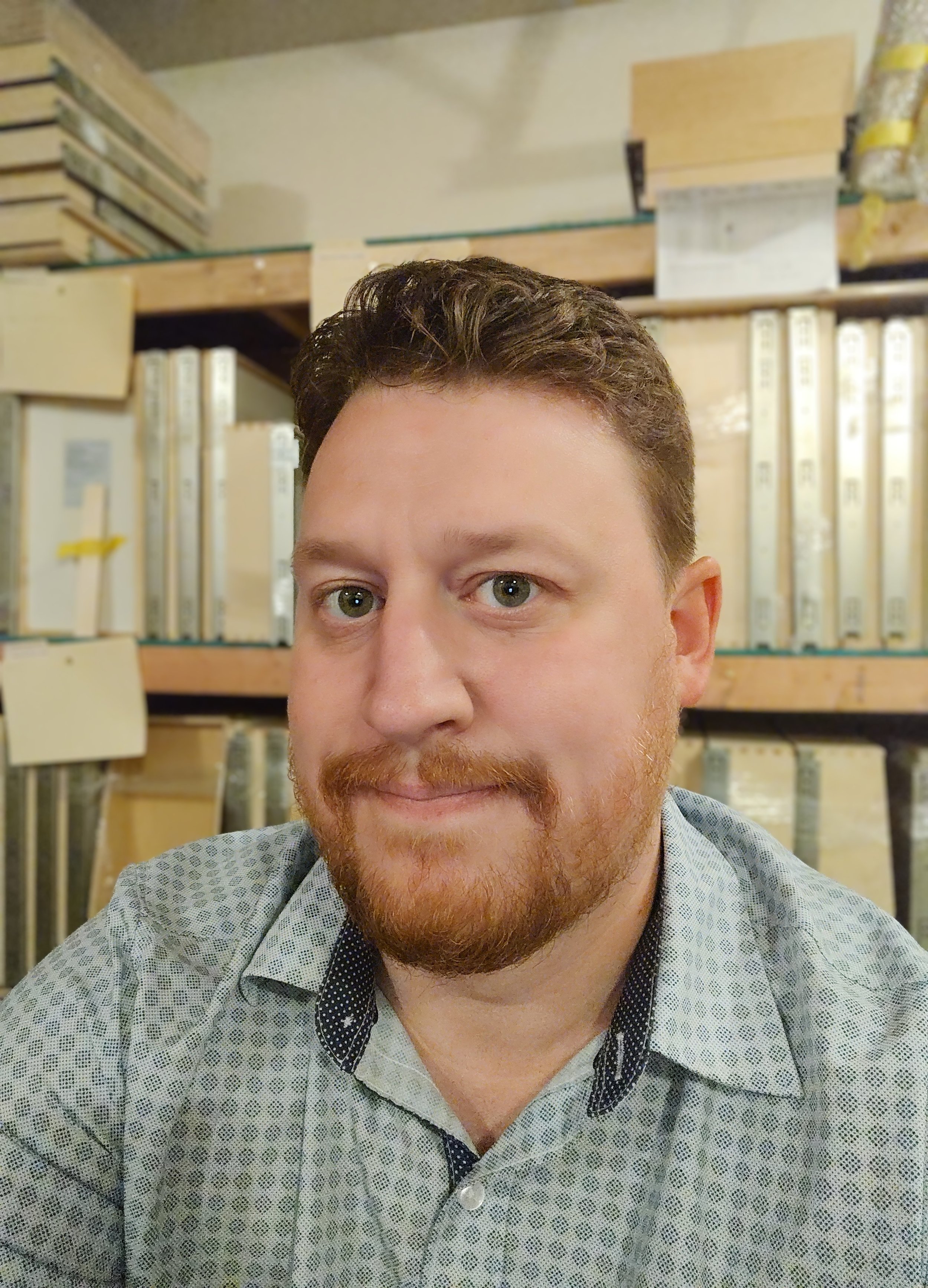 A man with brown hair and a beard taking a selfie in front of shelves with paint samples in a store.