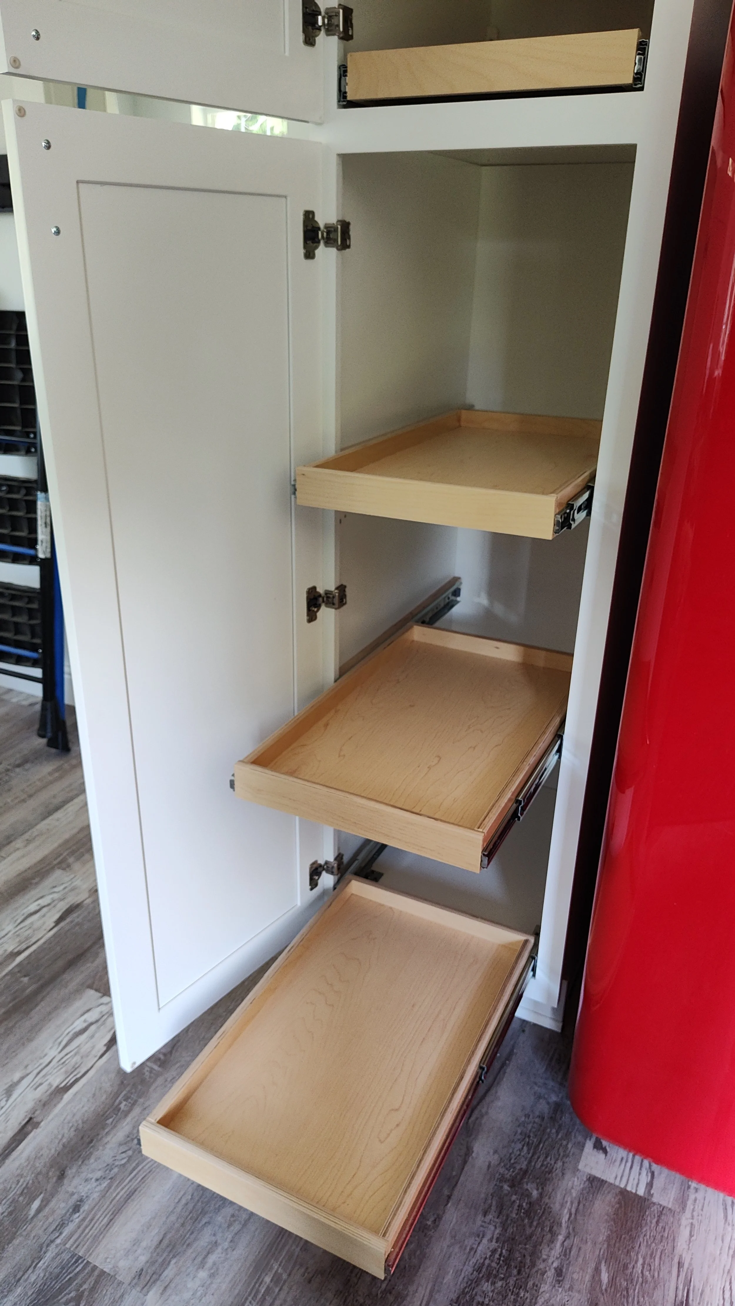 Open white cabinet with four empty wooden drawers partially pulled out, next to a red appliance or structure, on a wood floor.