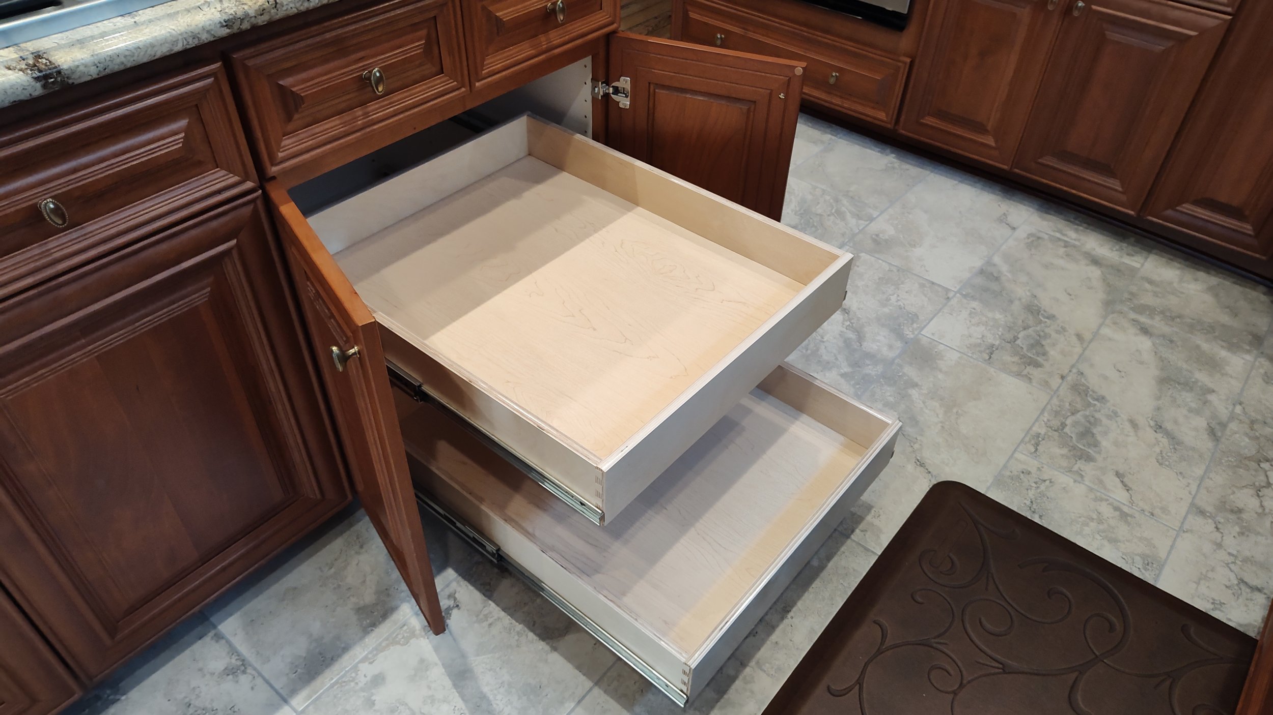 Open kitchen cabinet drawers with wooden interiors, dark brown cabinets, tiled floor, and a brown cushioned stool.