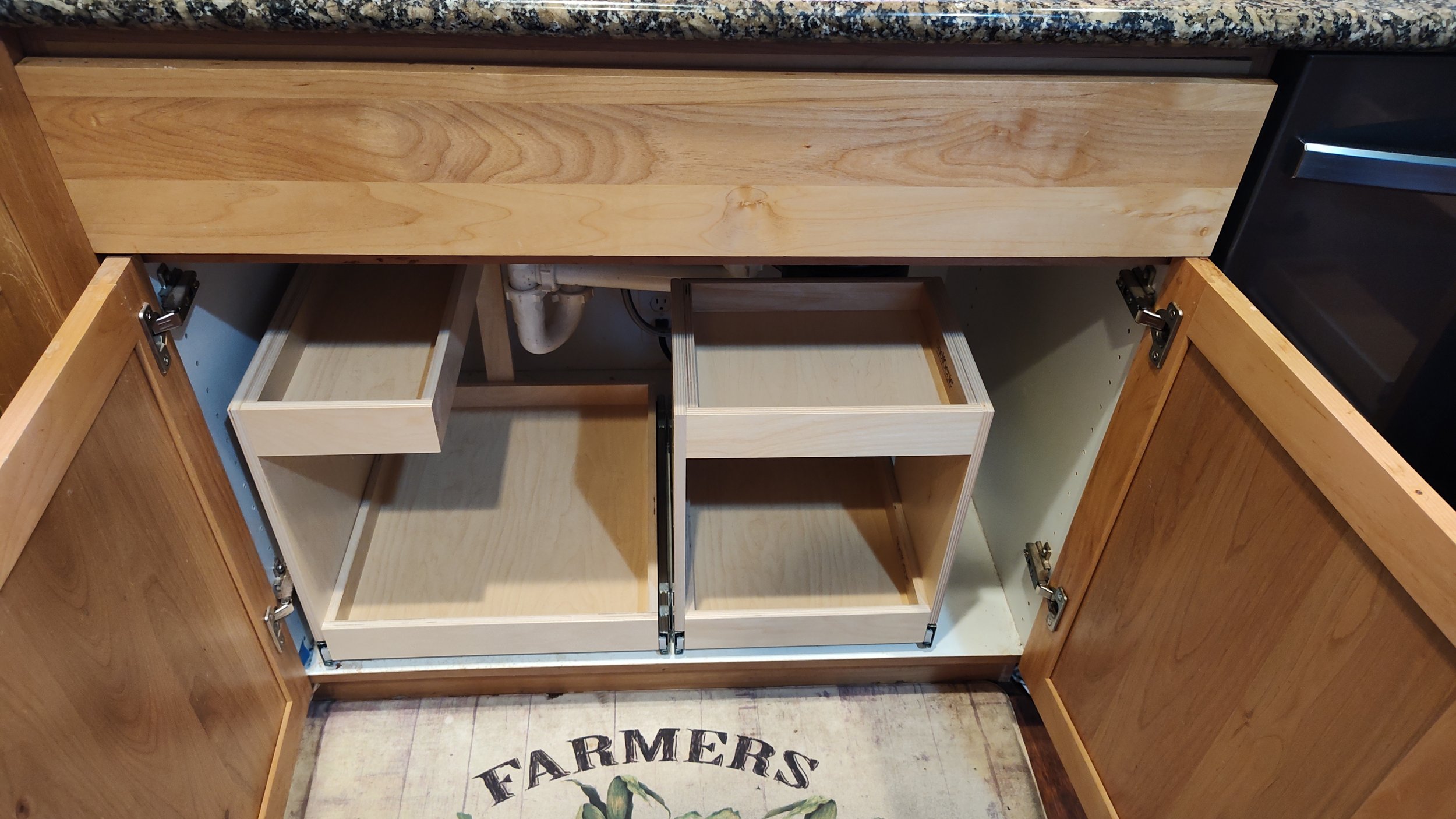 Open kitchen cabinet revealing unfinished wooden drawers and compartments with a granite countertop above.