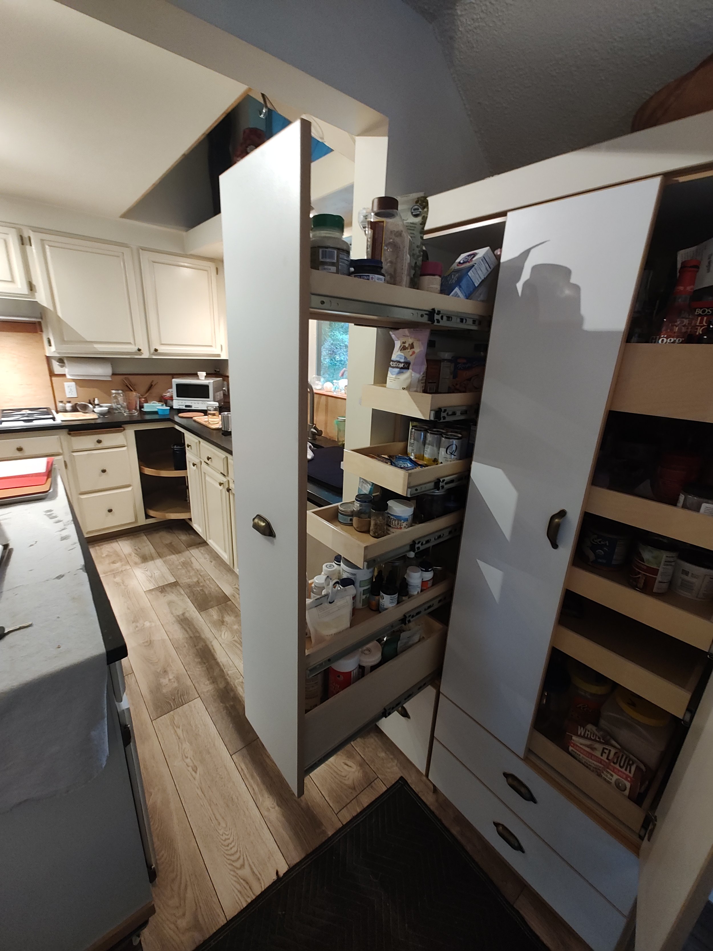 Open kitchen cabinets filled with spices, food containers, and pantry items in a kitchen with wooden flooring and cream-colored cabinets.