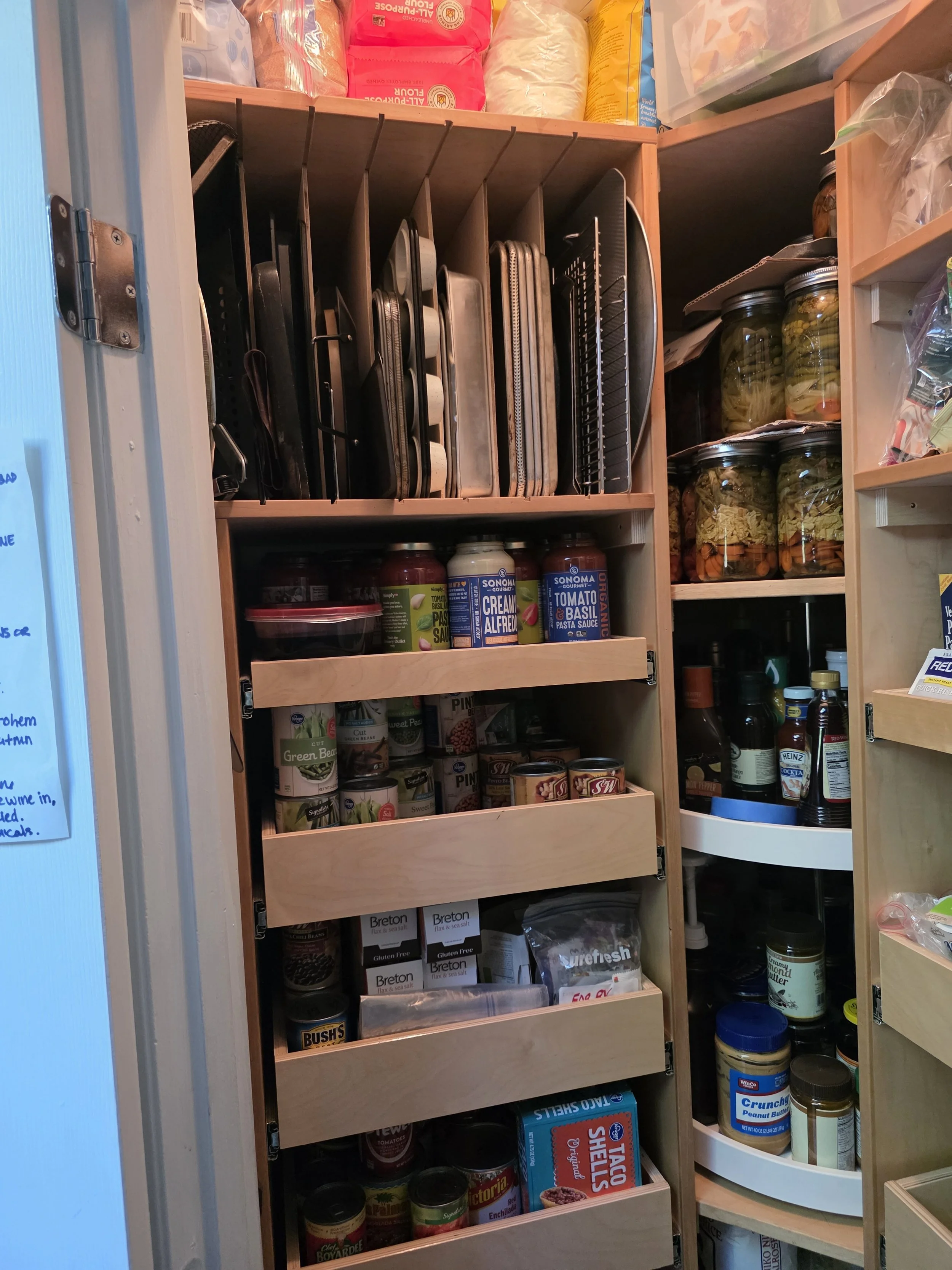 Pantry with shelves containing canned food, jars, boxed food, and kitchen supplies, with a section holding cutting boards and trays.