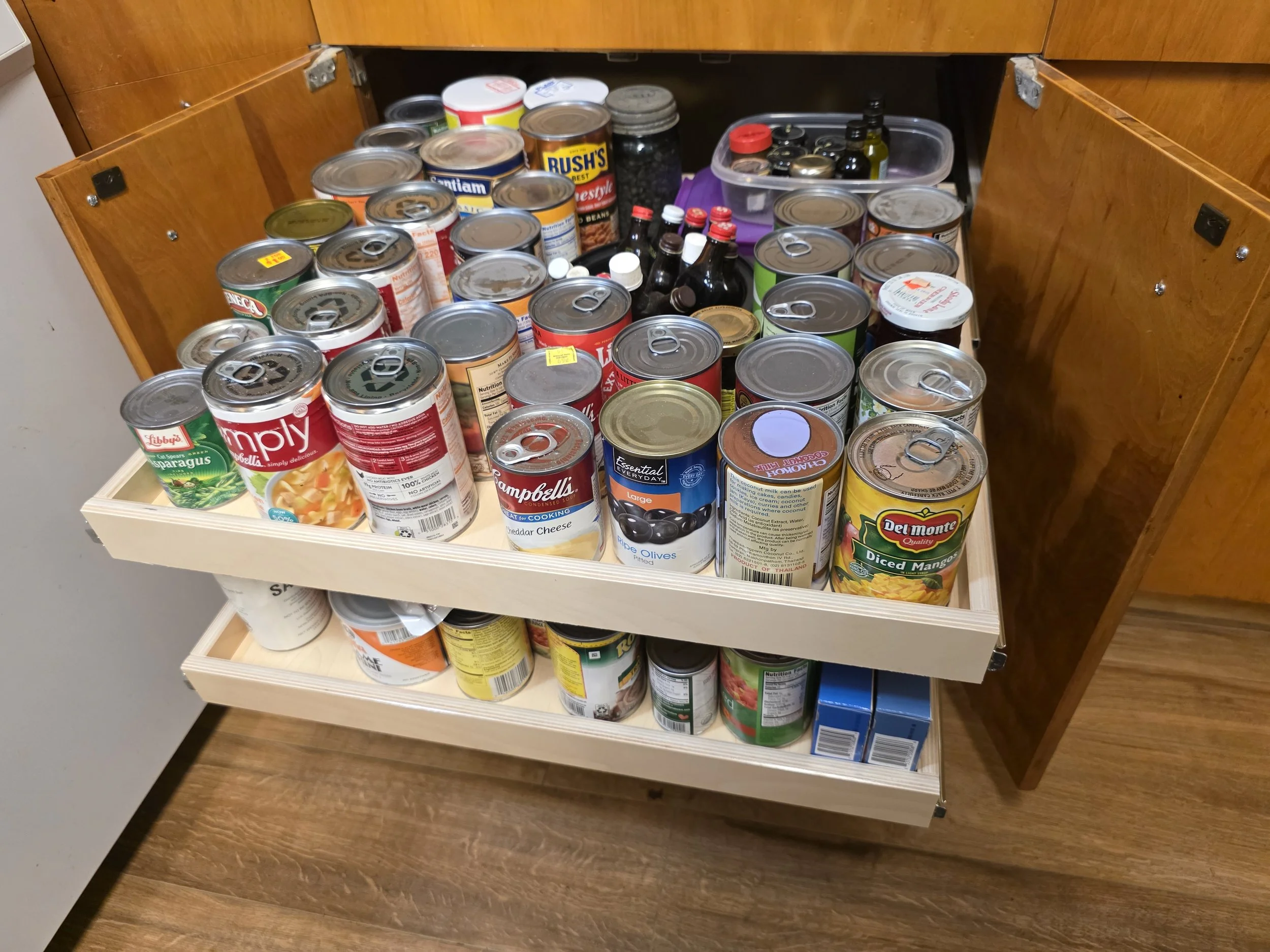 Open kitchen cabinet drawer filled with canned foods, jars, and bottles.