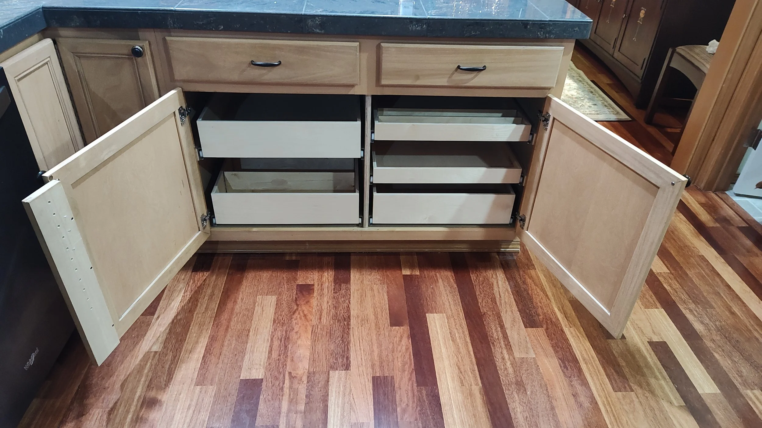 Open kitchen cabinet with empty drawers and doors, located beneath a black countertop with wooden cabinets on each side, hardwood floor.