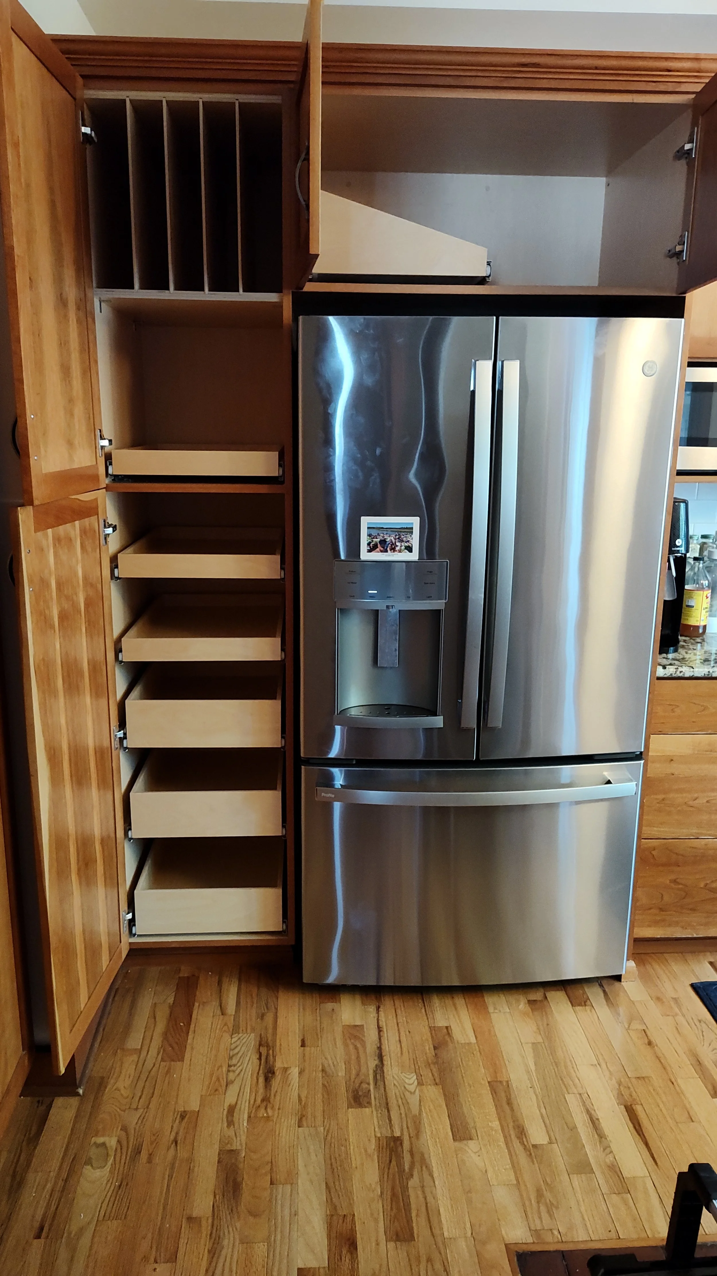 Stainless steel refrigerator with a water and ice dispenser, positioned beside open kitchen cabinets with pull-out shelves and wine storage compartments, and wooden kitchen furniture.