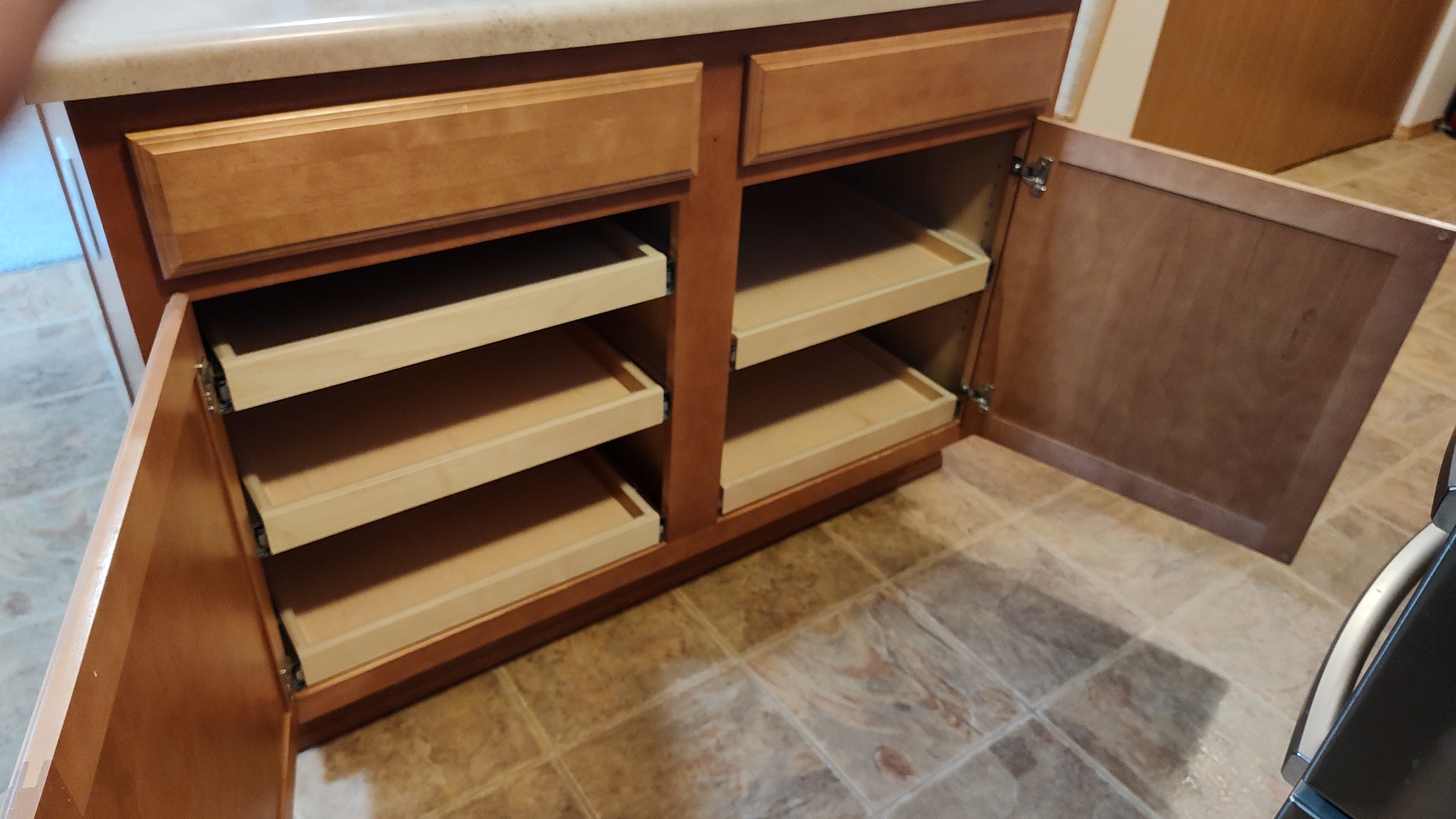 Open kitchen cabinet with empty pull-out drawers, wooden finish, in a tiled floor kitchen.