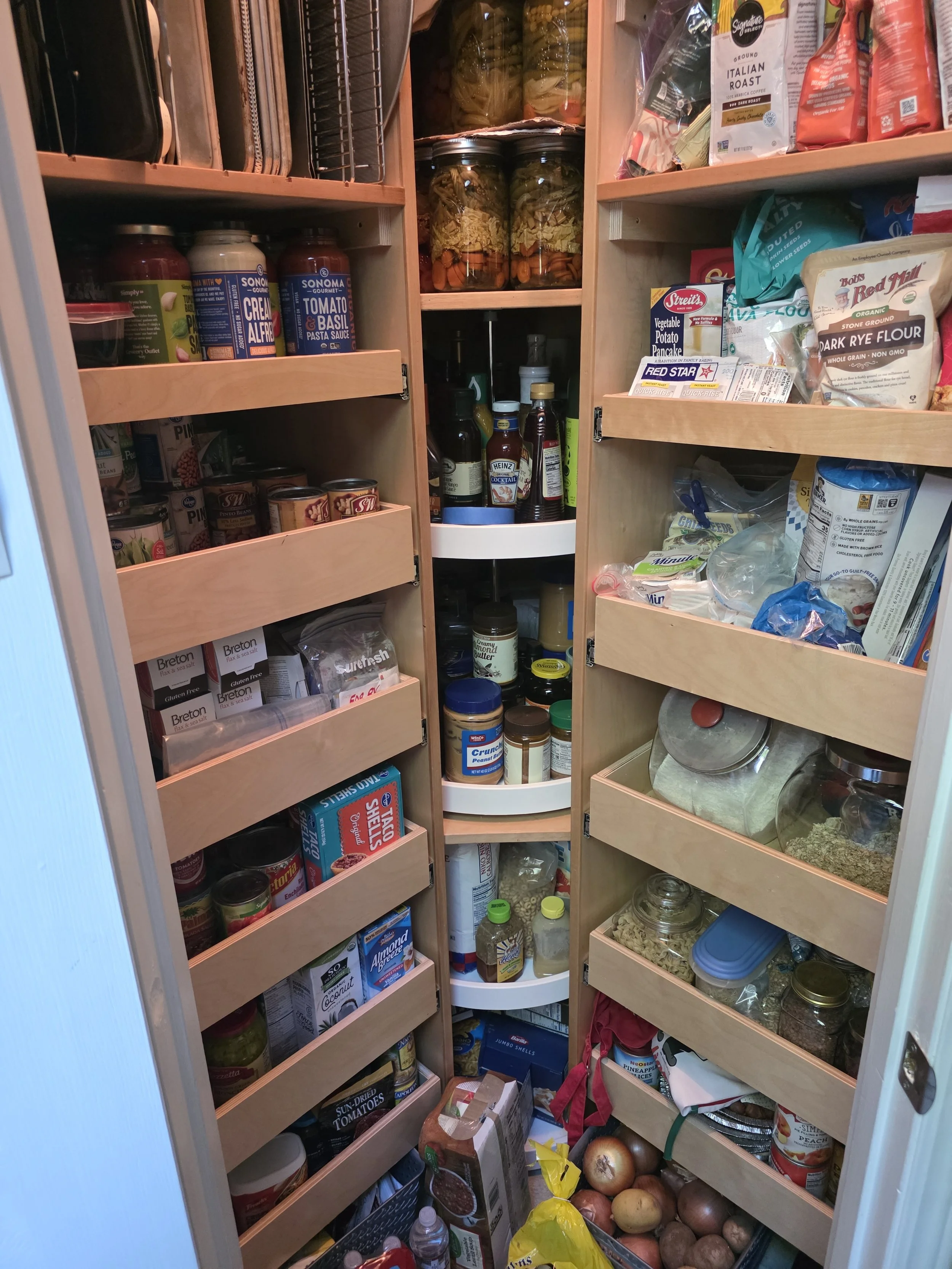 Full pantry shelf filled with various canned goods, jars, boxes, bottles, and food items, including pasta, sauces, spices, vegetables, and onions on the floor.