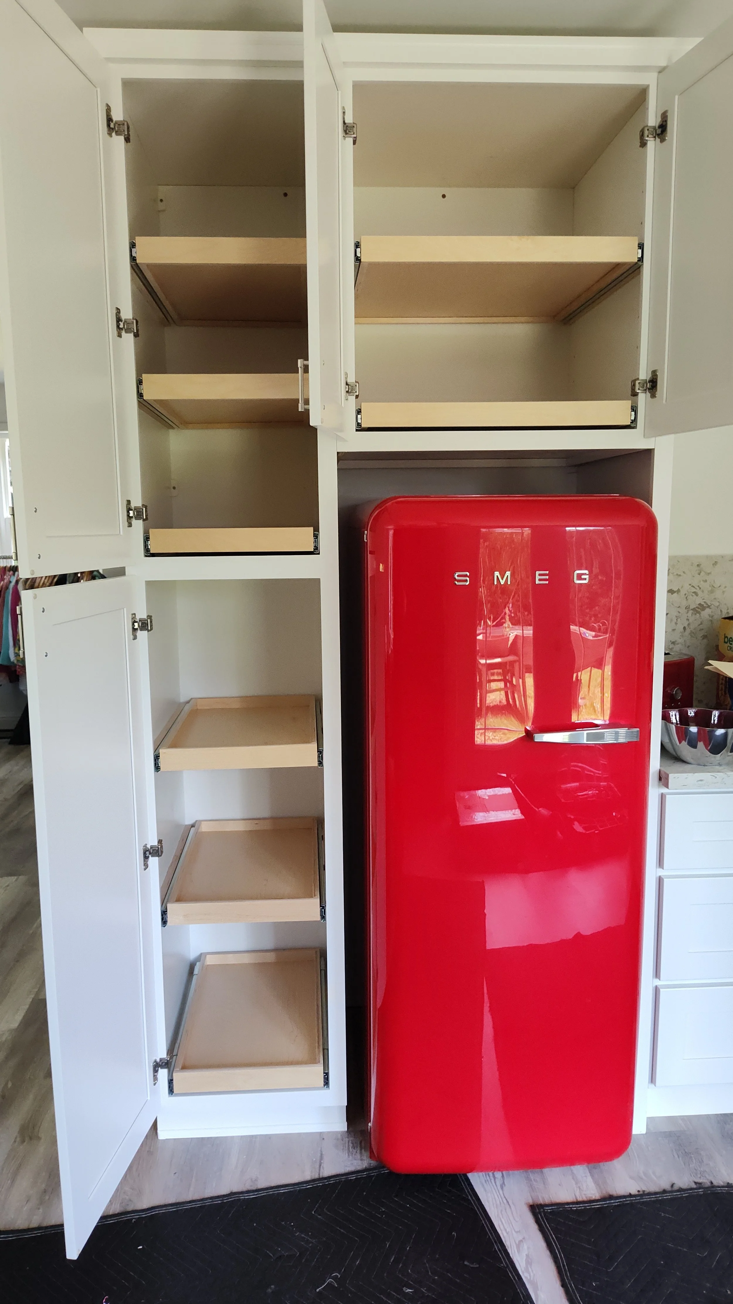Empty white kitchen cabinet with open doors and pull-out shelves next to a red Smeg refrigerator.