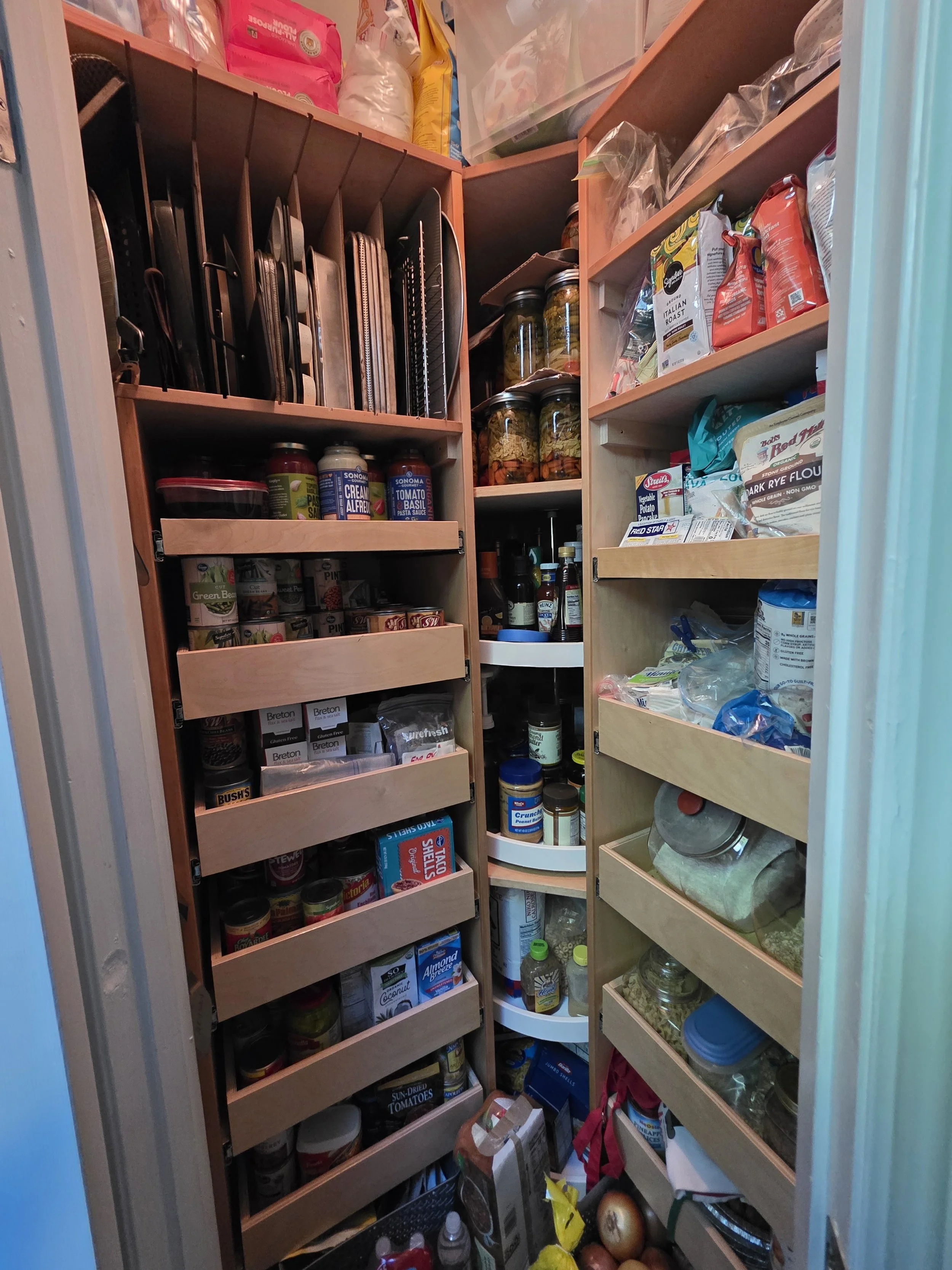 A small pantry with shelves filled with canned goods, jars, bags of food, and kitchen supplies, organized but densely packed.
