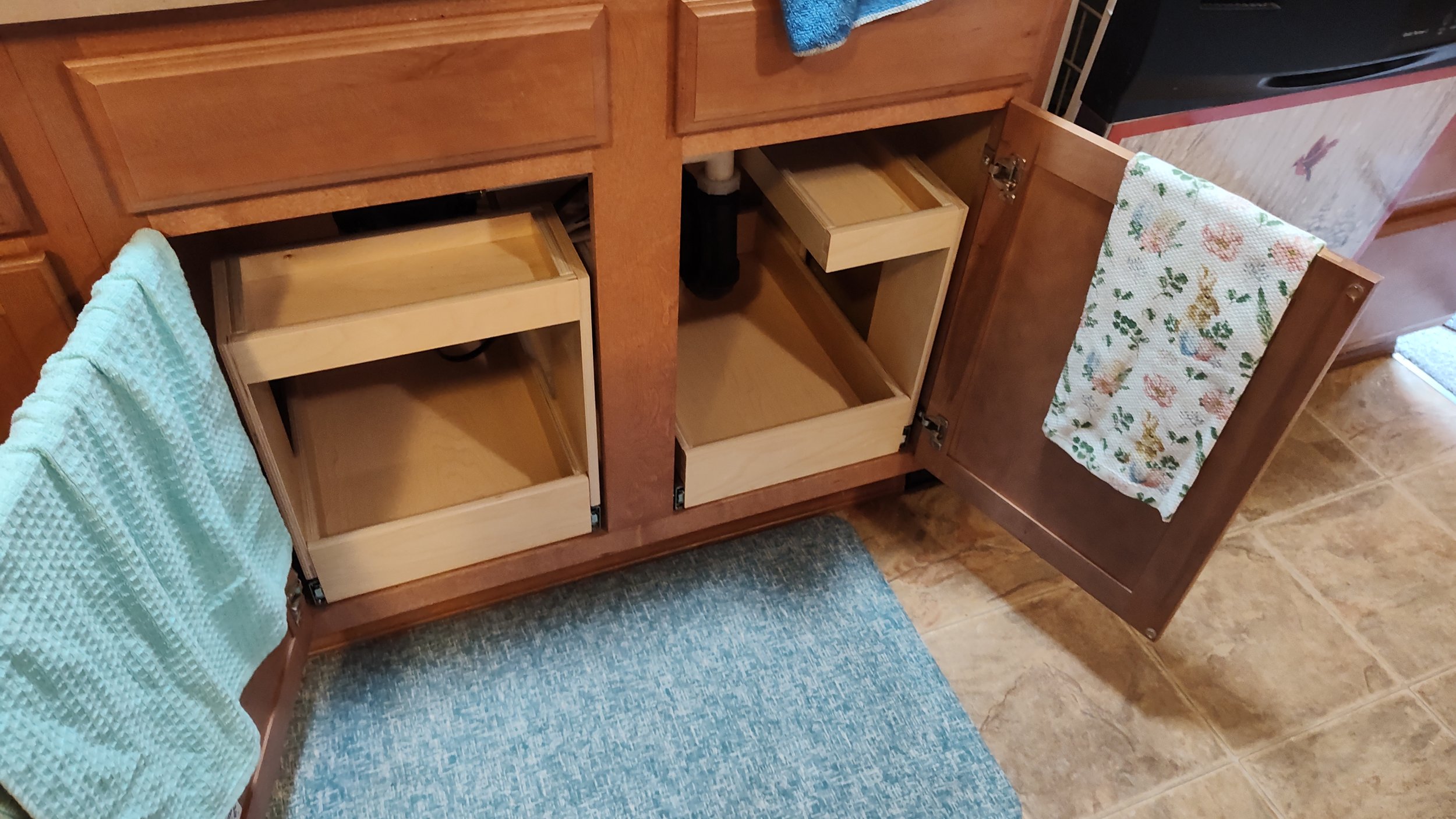 Open kitchen cabinet with two empty pull-out drawers and a door with a decorated towel hanging on it. Nearby is a blue cushioned stool and beige tiled floor.