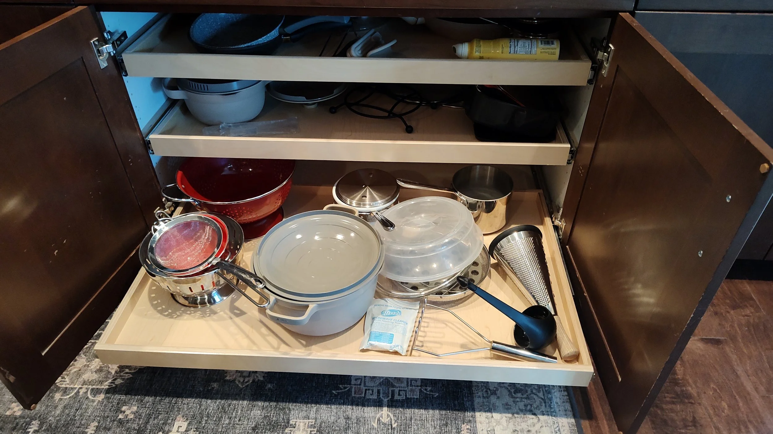 Open kitchen cabinet drawer filled with various cookware including pots, pans, strainers, and utensils.